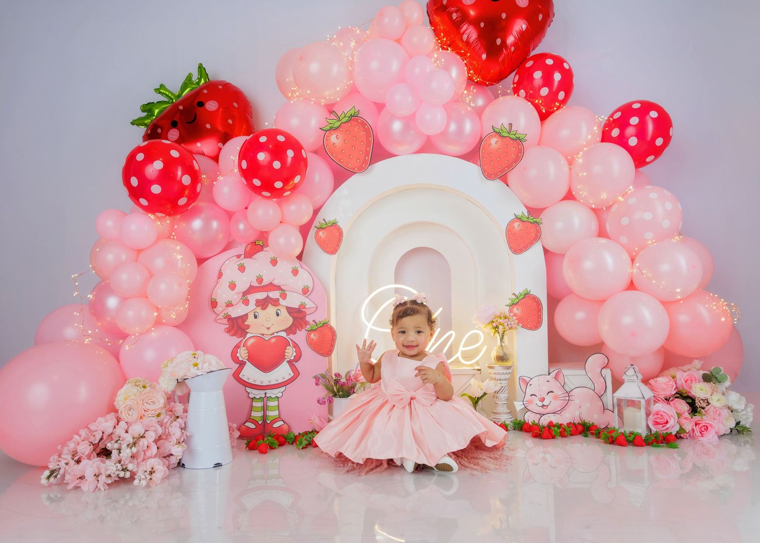 Baby girl in pink dress sits before a strawberry-themed birthday backdrop with balloons and decorations.