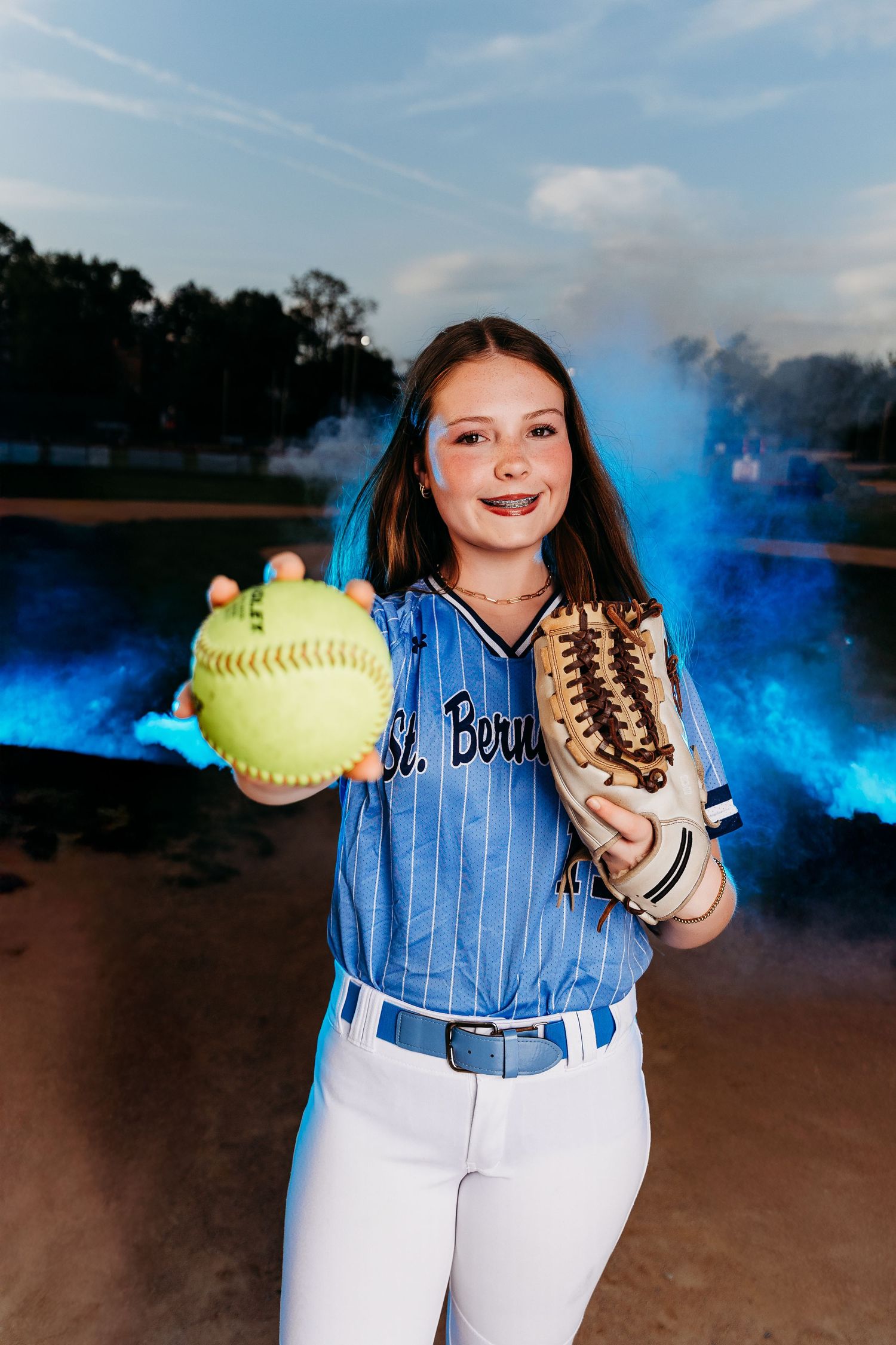 Young softball player in blue St. Bernard jersey holds yellow ball and glove with blue smoke in background.