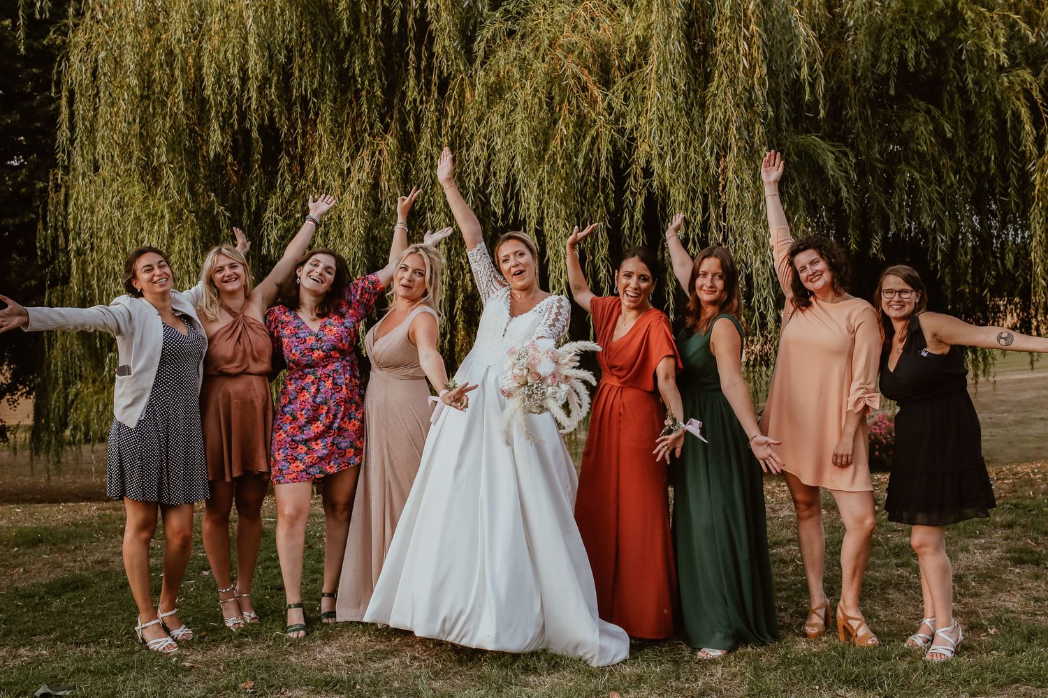Bride in white gown celebrates with bridesmaids in colorful dresses under a willow tree outdoors.