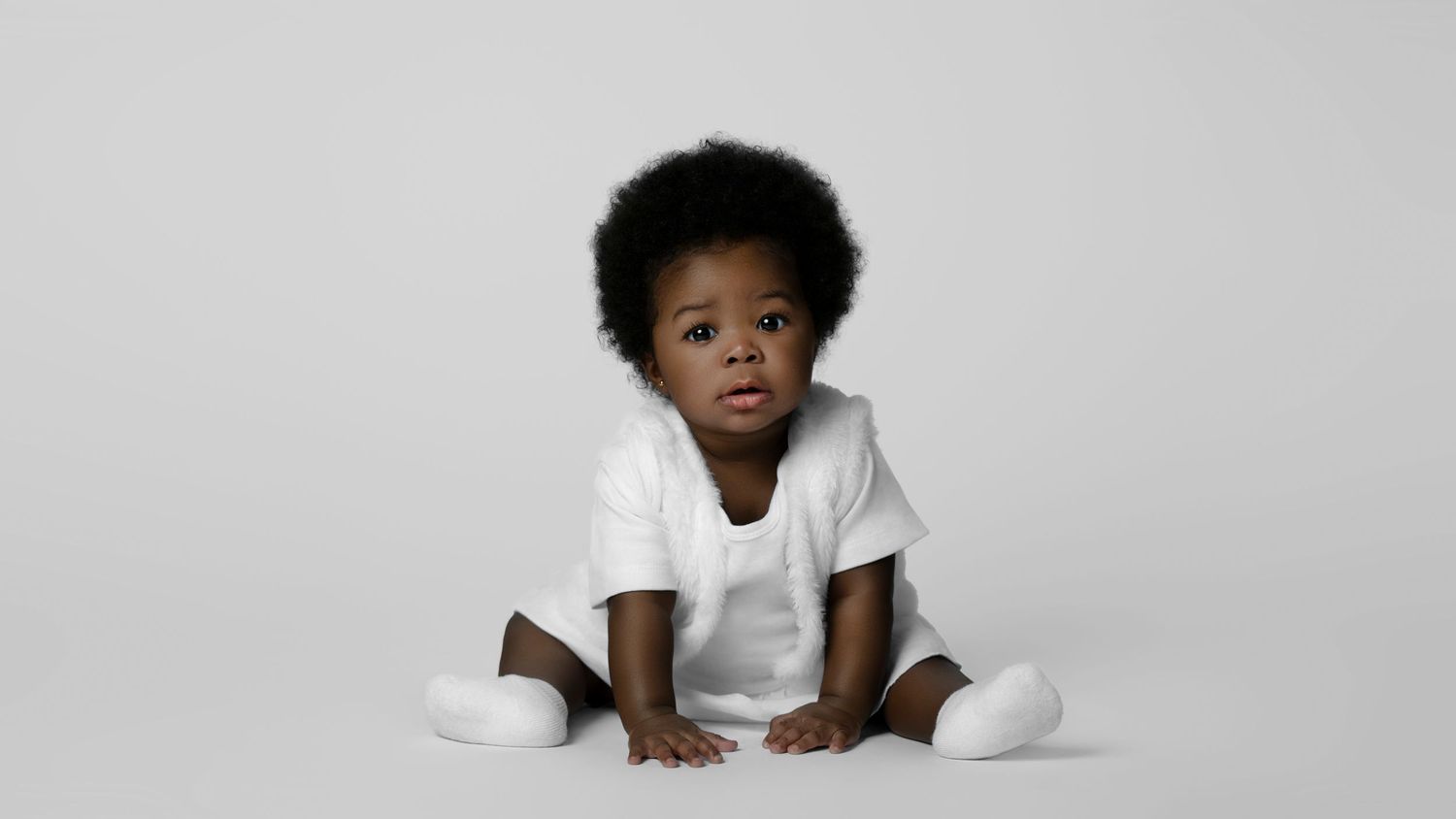 Adorable Black baby in white outfit sitting on white background, looking up curiously.