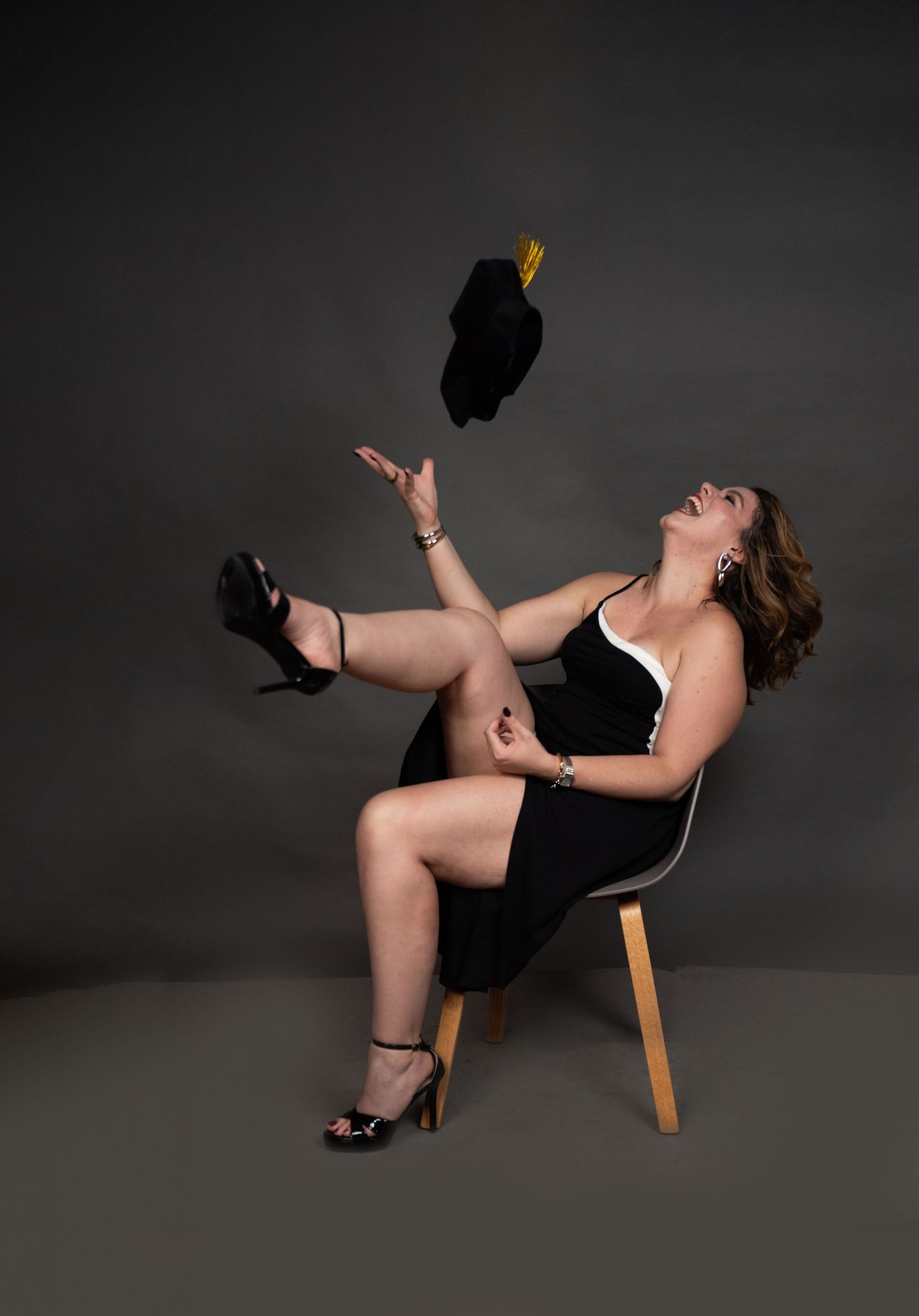 Woman in black dress tosses graduation cap while seated on modern chair, celebrating academic achievement.