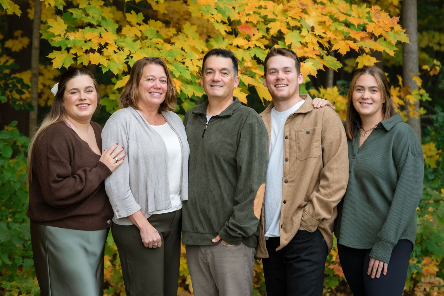 Five people smiling together outdoors in front of vibrant autumn foliage in shades of yellow, orange, and green.