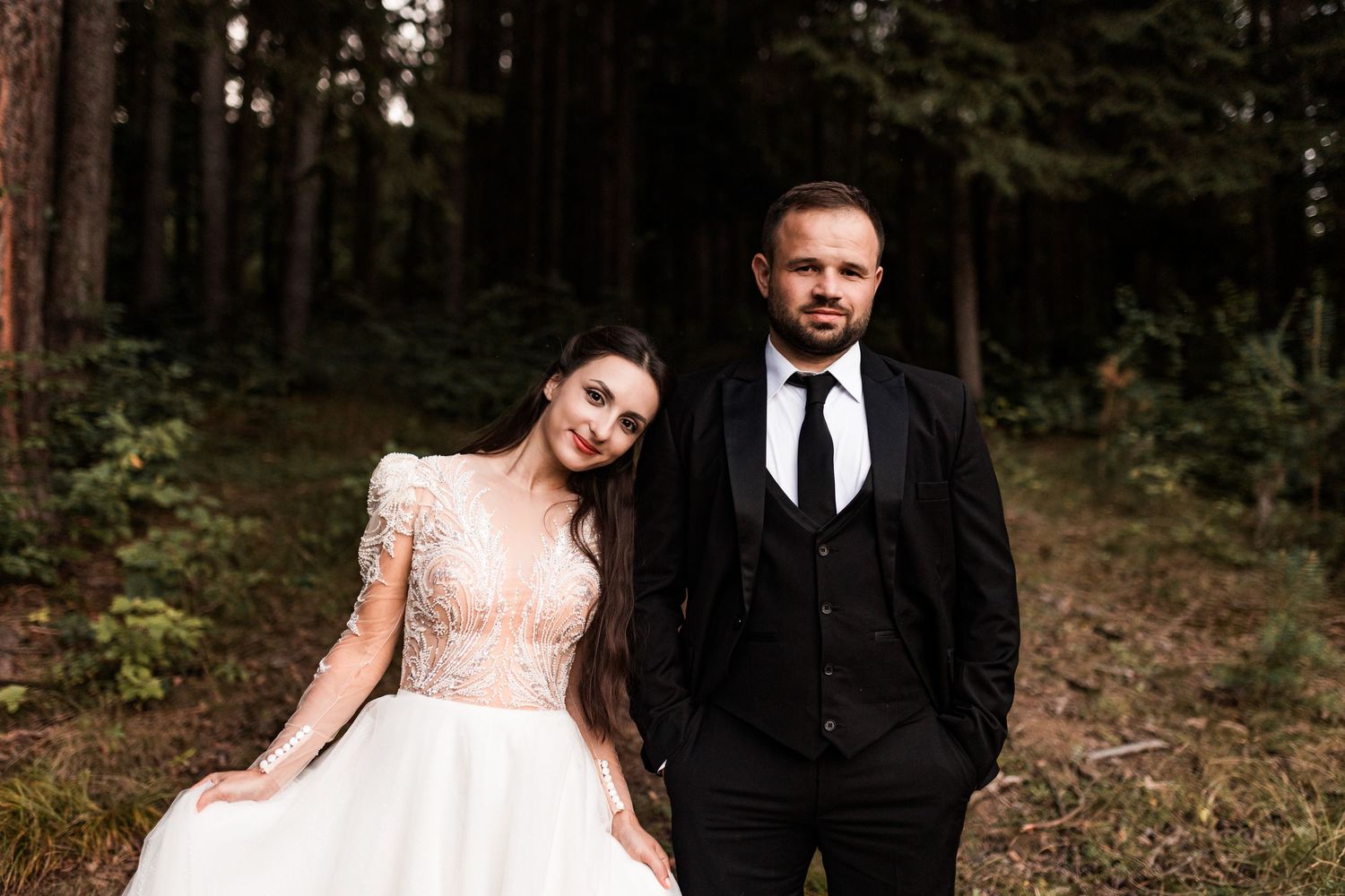 Bride and groom posing in a forest, woman in white gown, man in black suit with tie, wedding photography.