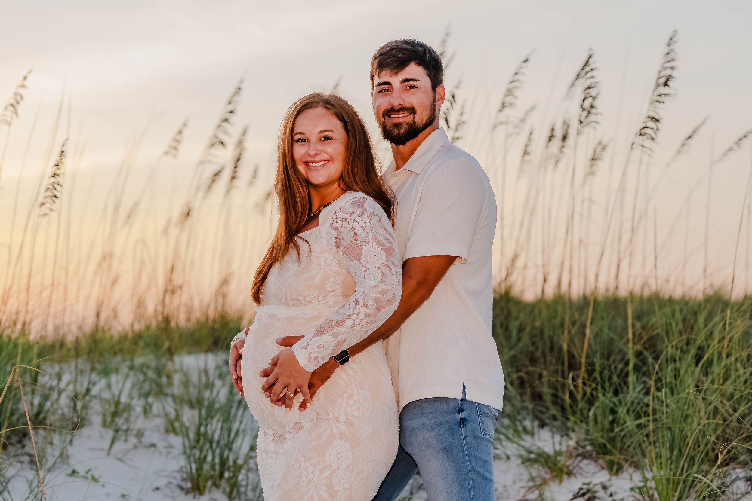 Pregnant couple posing on sandy beach at sunset with sea oats, woman in lace maternity gown, man in white shirt.
