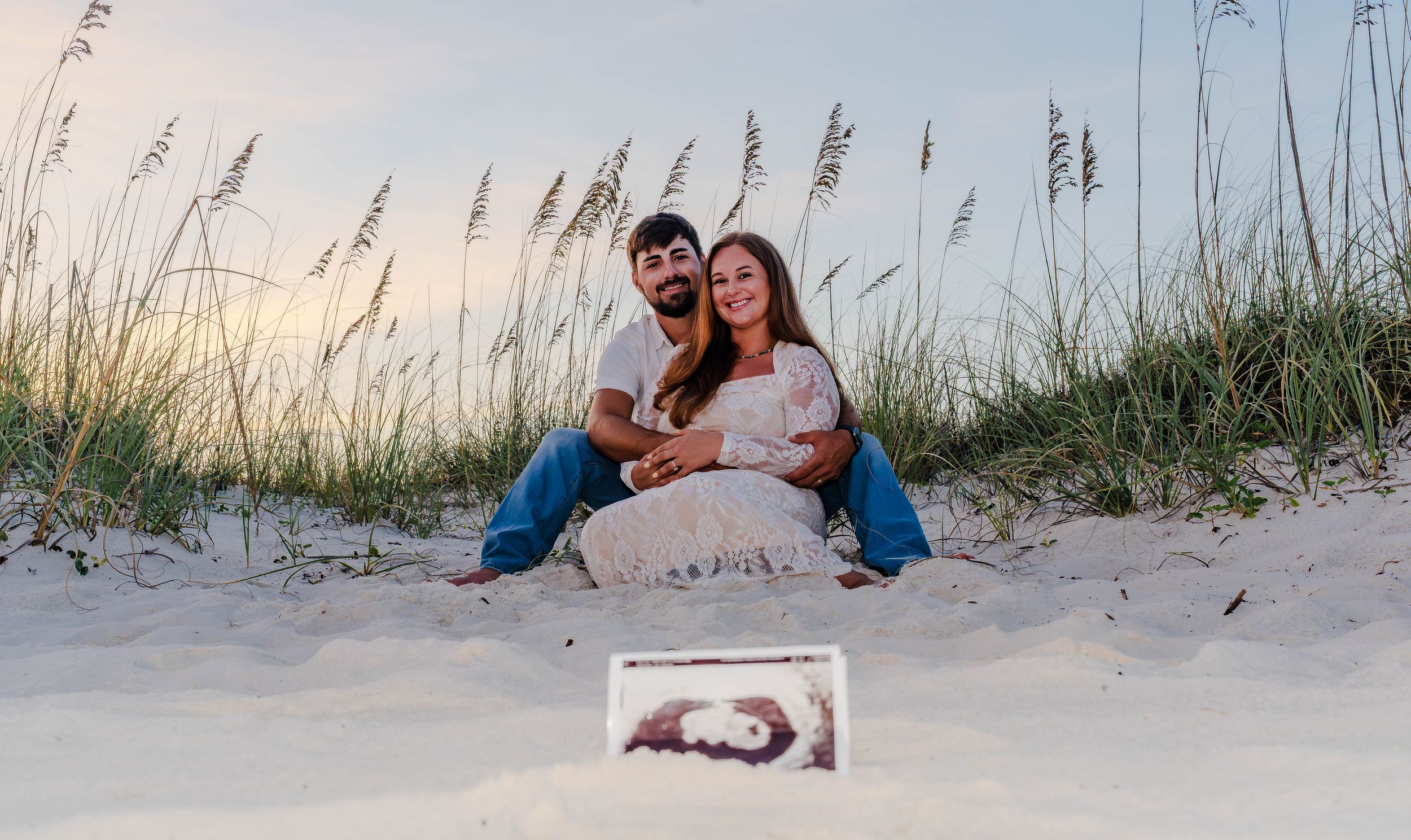 Couple sitting on beach sand dunes at sunset with ultrasound photos displayed in front, maternity announcement photo.