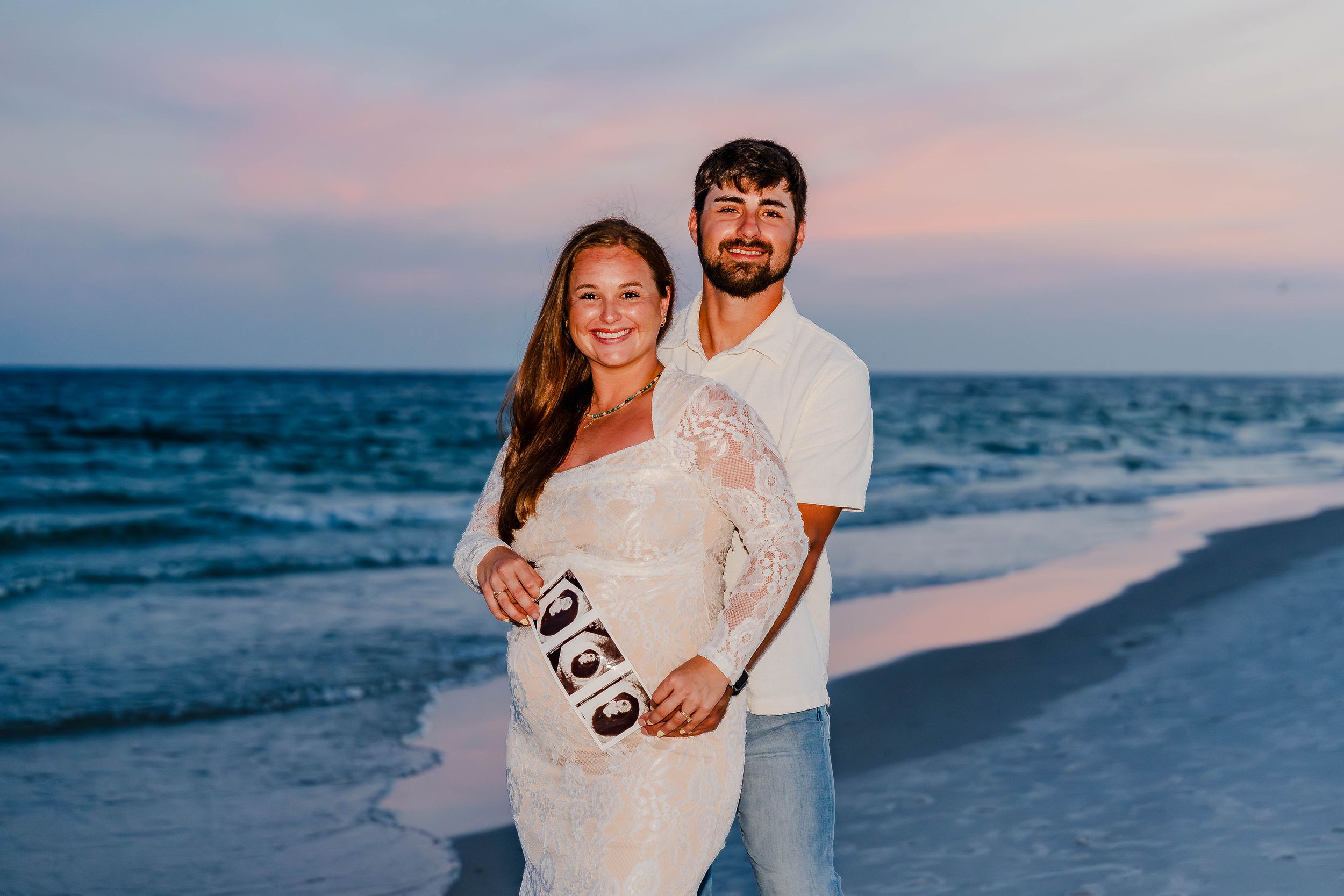 Pregnant couple holding ultrasound photos on a beach at sunset, woman in white lace dress, man in white shirt.
