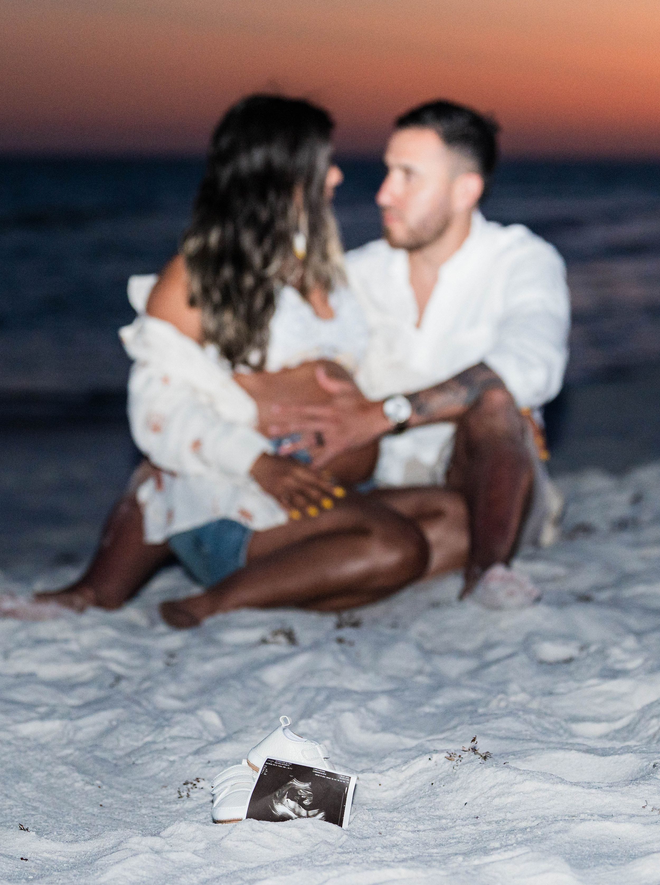 Couple sitting on beach at sunset with ultrasound photo in foreground, pregnancy announcement.