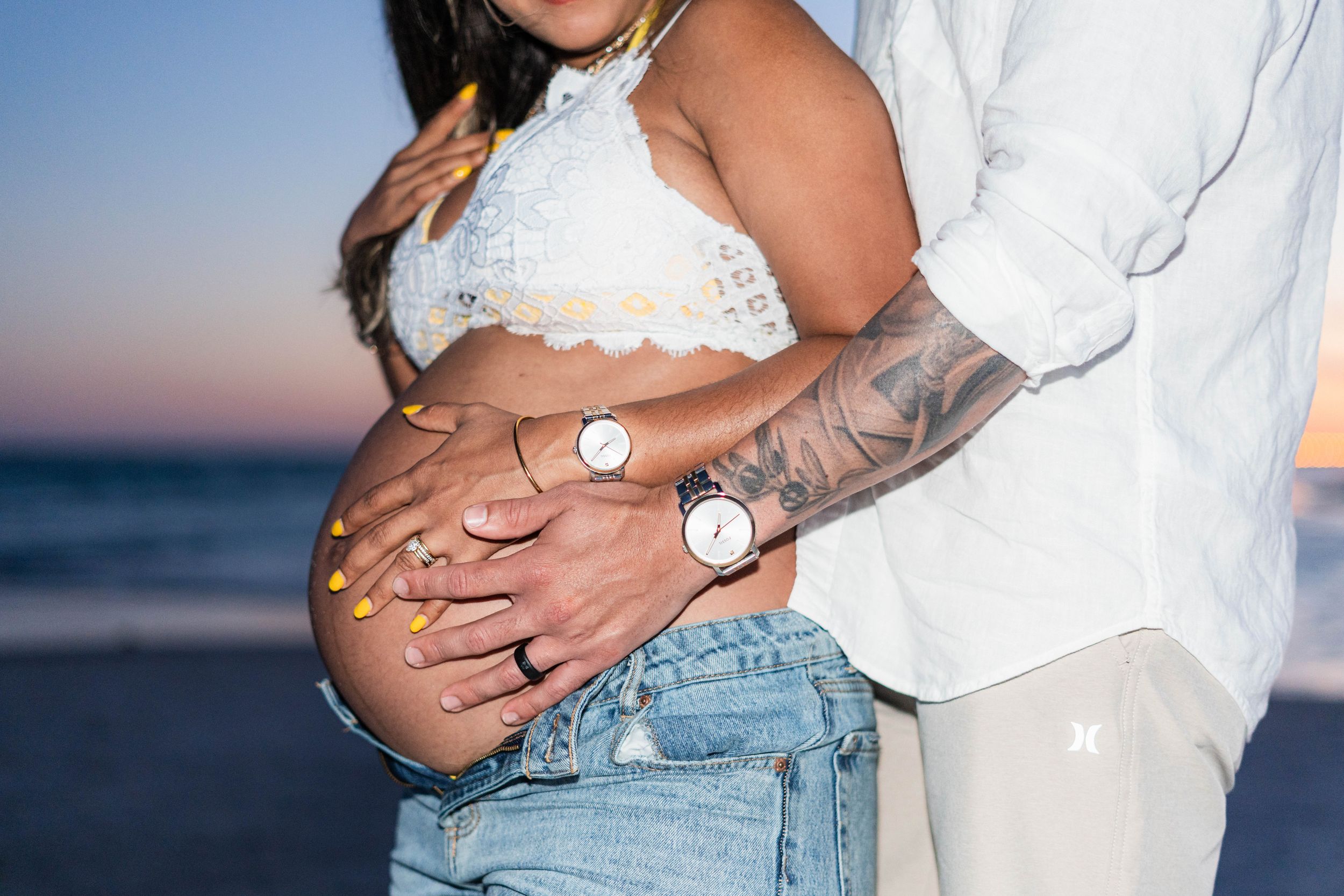 Pregnant couple at beach sunset, woman in crop top and jeans with tattooed partner embracing her baby bump.