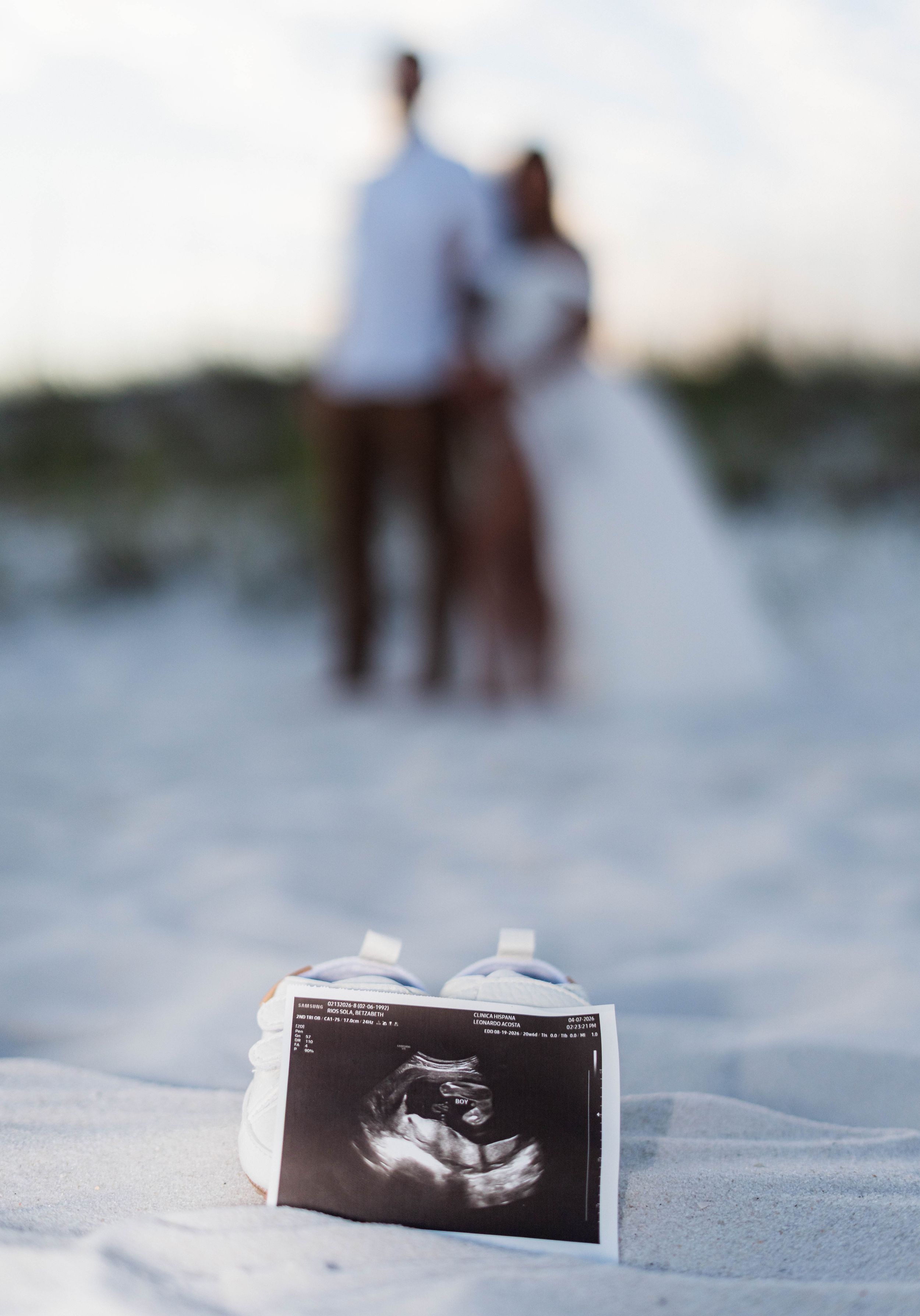 Expecting couple stands on white sand beach behind ultrasound photo and tiny baby shoes announcement.