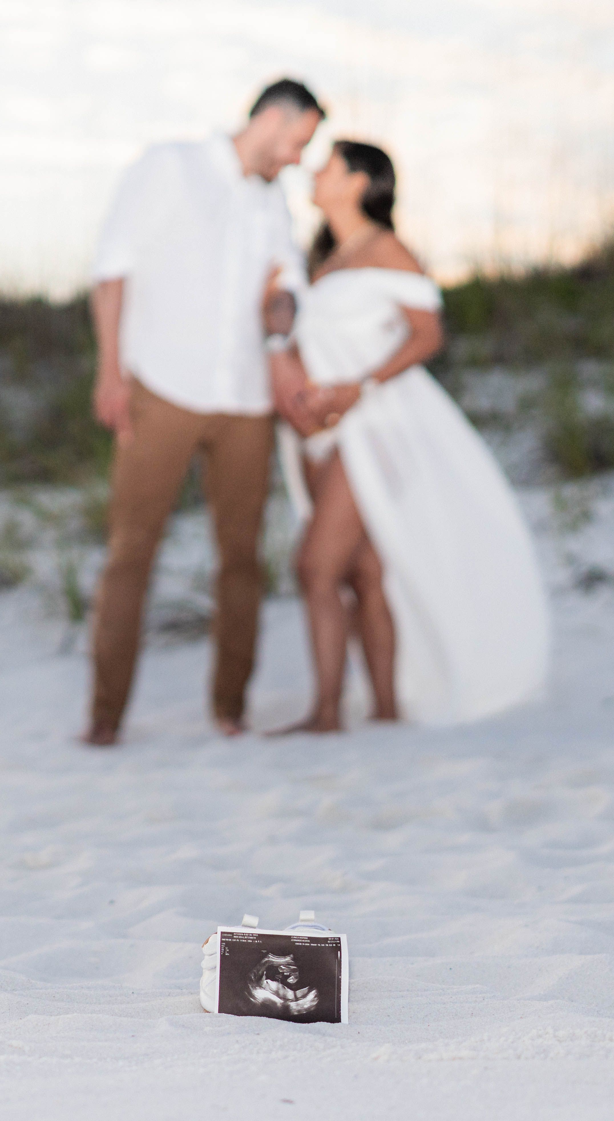 Couple in white outfits on sandy beach with ultrasound photo in foreground for maternity announcement.