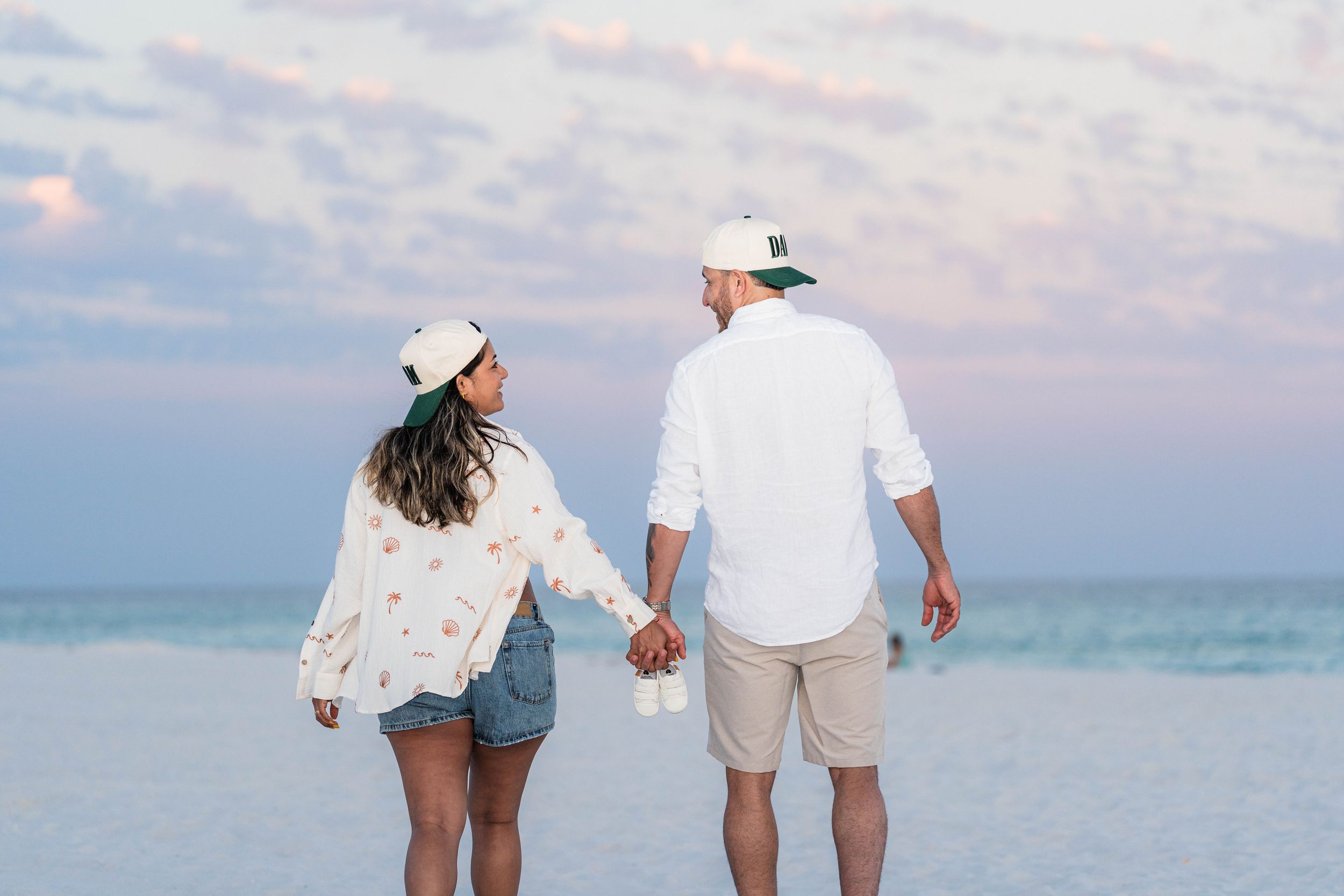 Couple holding hands on a beach at sunset, wearing white outfits and caps, with calm ocean waves behind them.