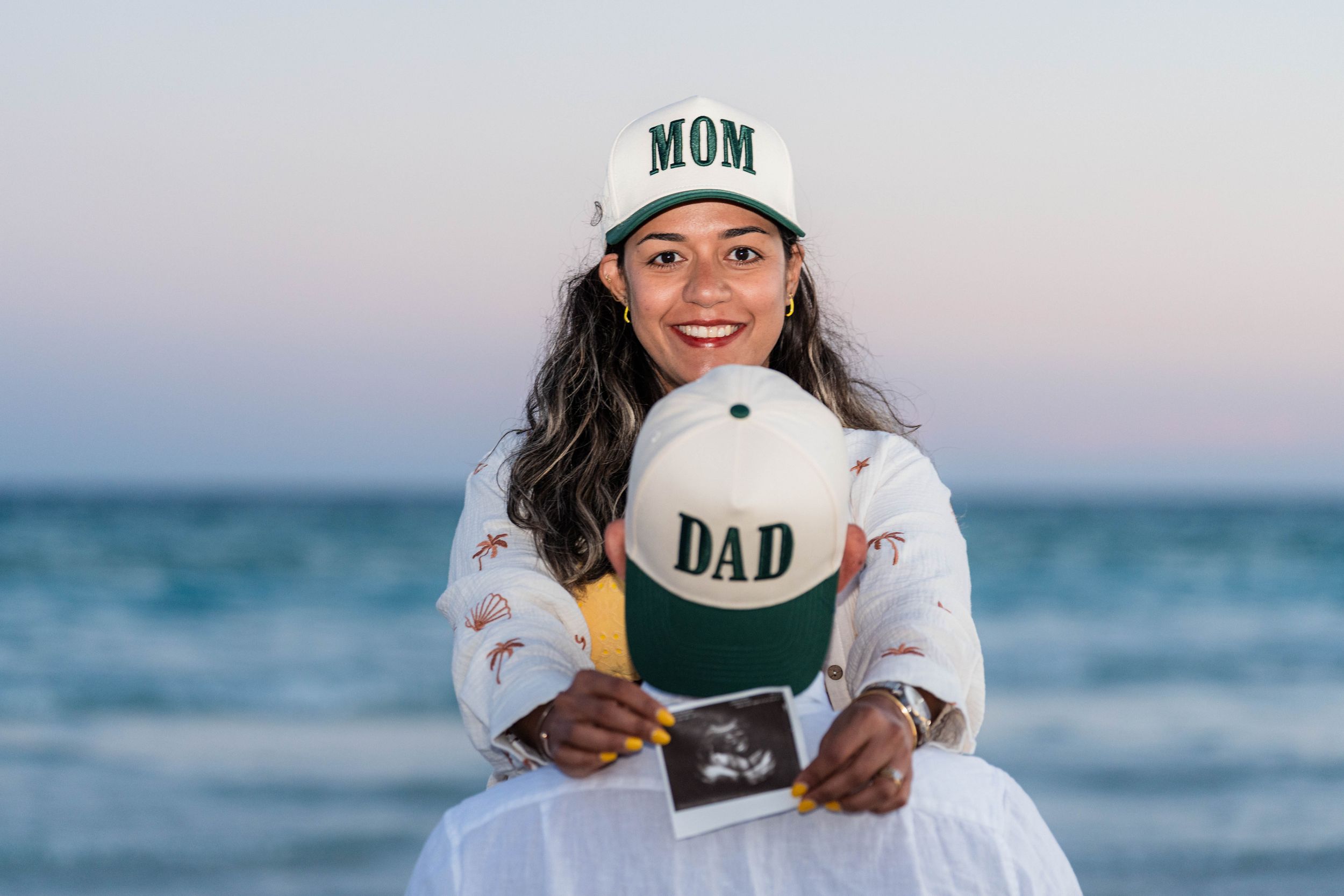 Pregnant woman at beach holding MOM and DAD hats with ultrasound photo, announcing pregnancy.