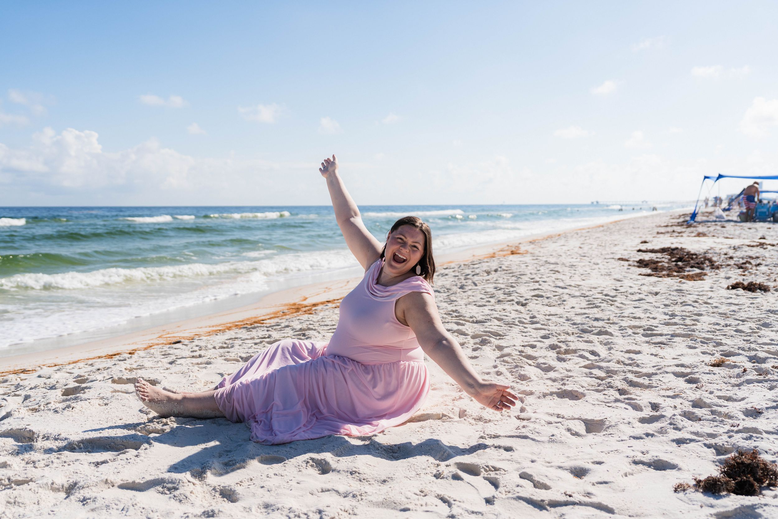 A joyful woman sits on a white sandy beach, raising her fist in celebration with ocean waves behind her.