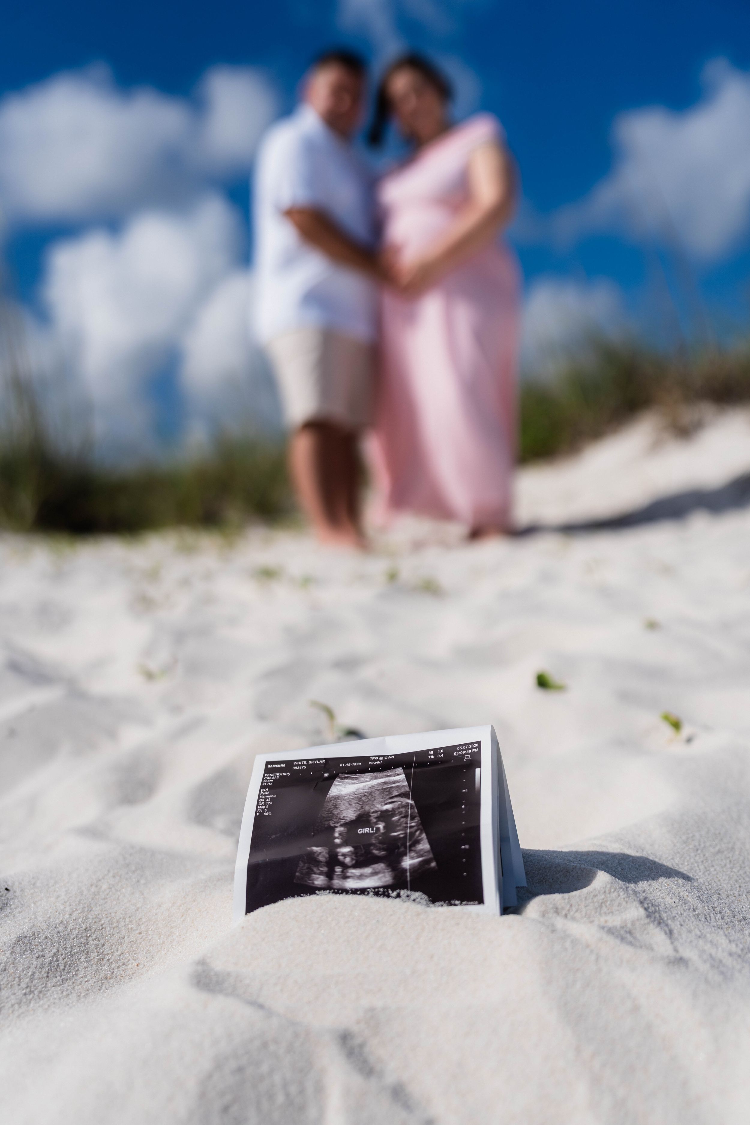 Ultrasound photo in sand with pregnant couple embracing on sunny beach in background.