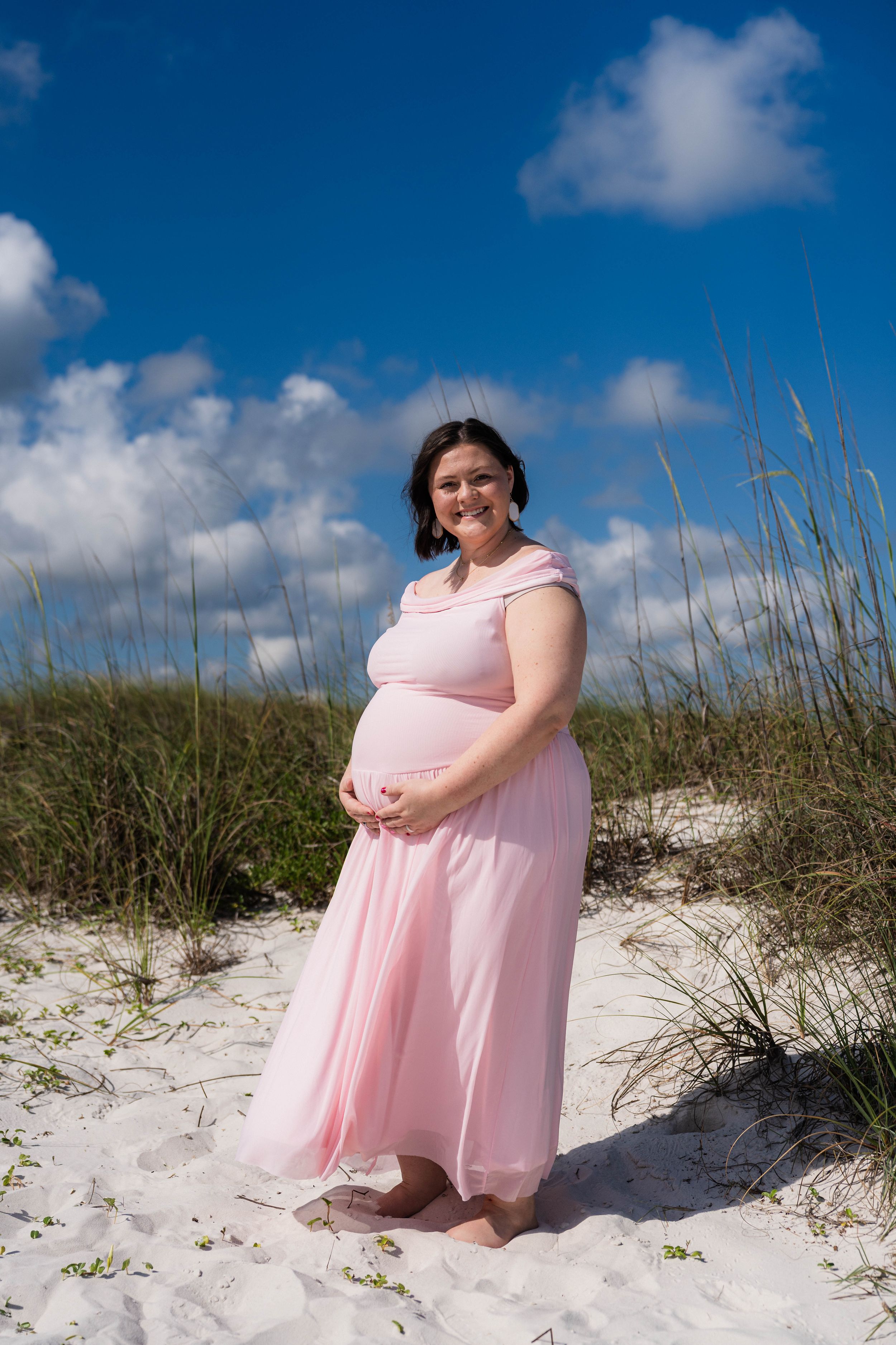Pregnant woman in pink dress holding baby bump on white sand beach with blue sky and sea oats.