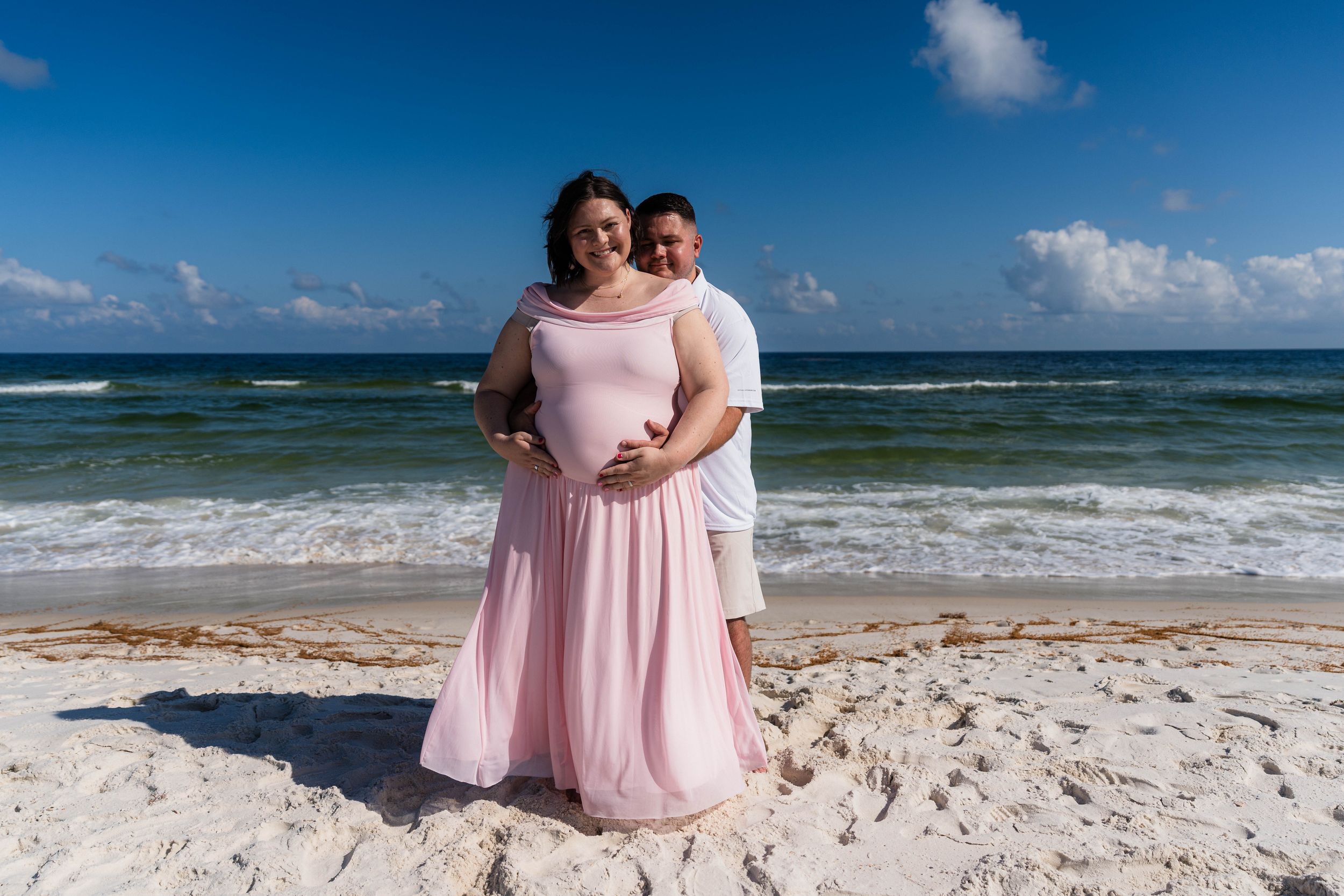 Couple poses on white sand beach, woman in pink gown with partner embracing her bump, ocean waves behind them.