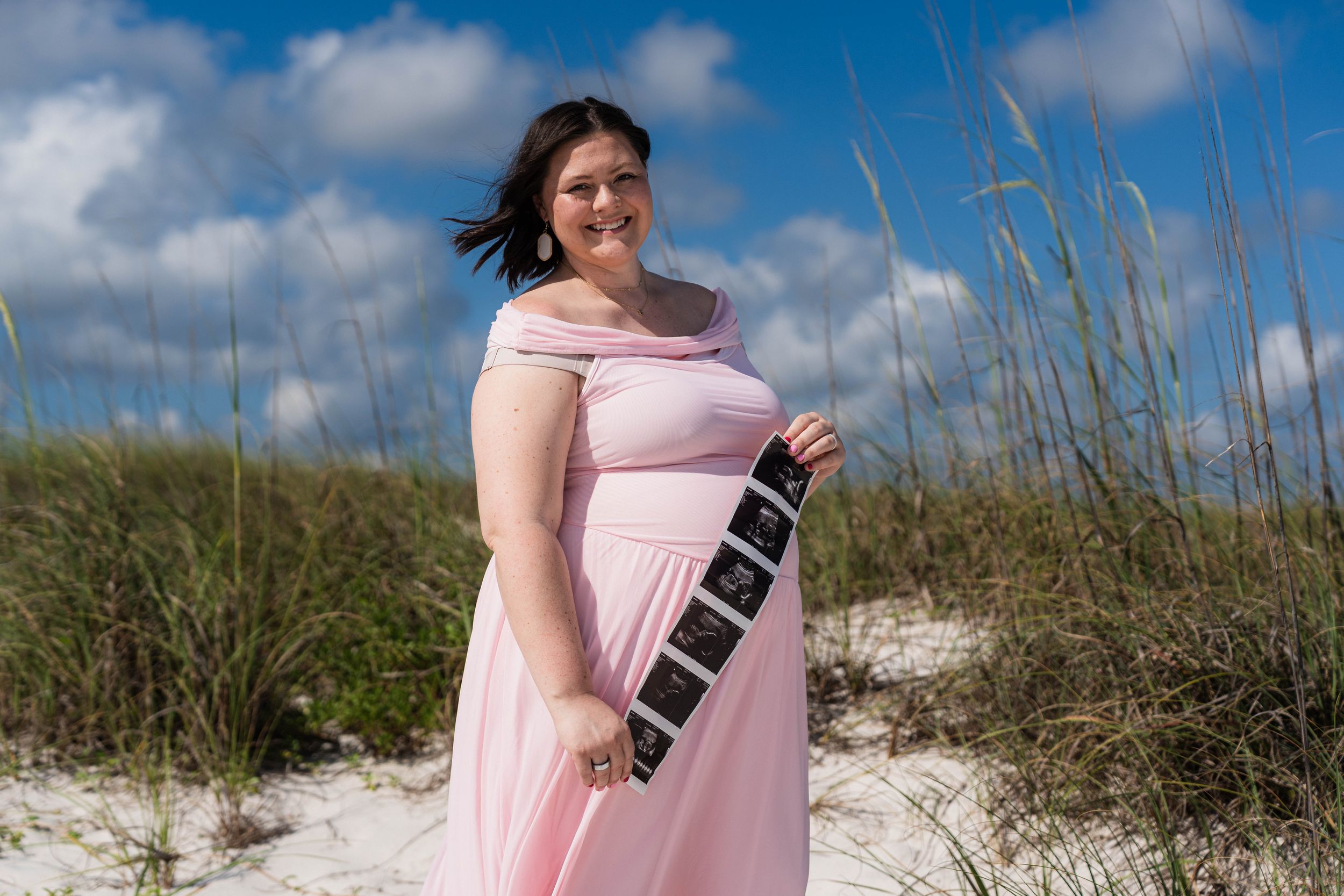 Pregnant woman in pink dress holding ultrasound photos on sandy beach with sea oats and blue sky.