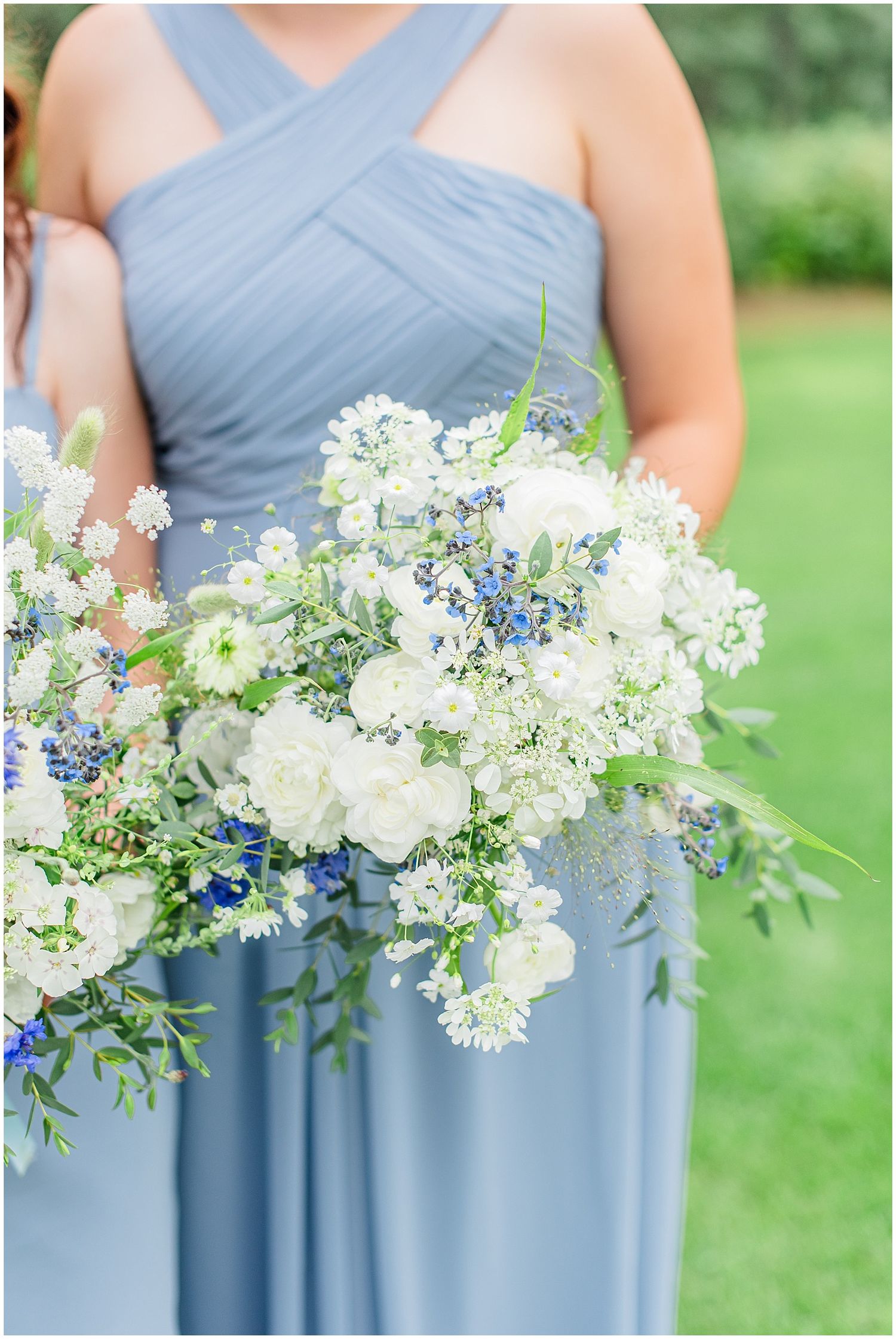 Bridesmaid in dusty blue dress holding lush white and blue floral bouquet with greenery outdoors.