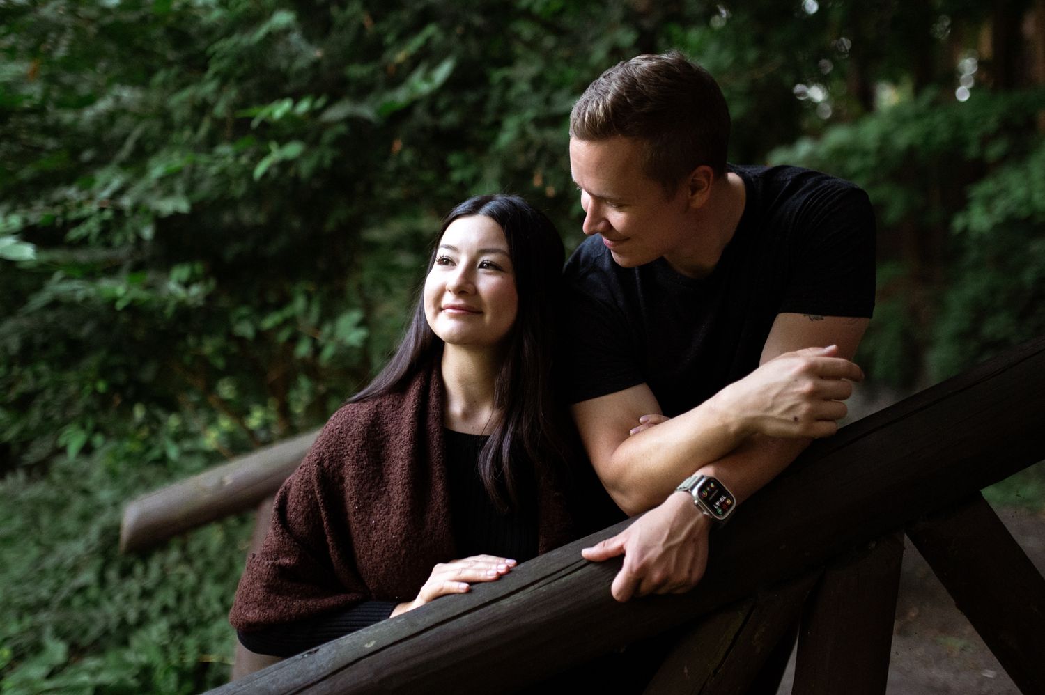 A smiling couple sharing a tender moment on a wooden deck surrounded by lush green trees.