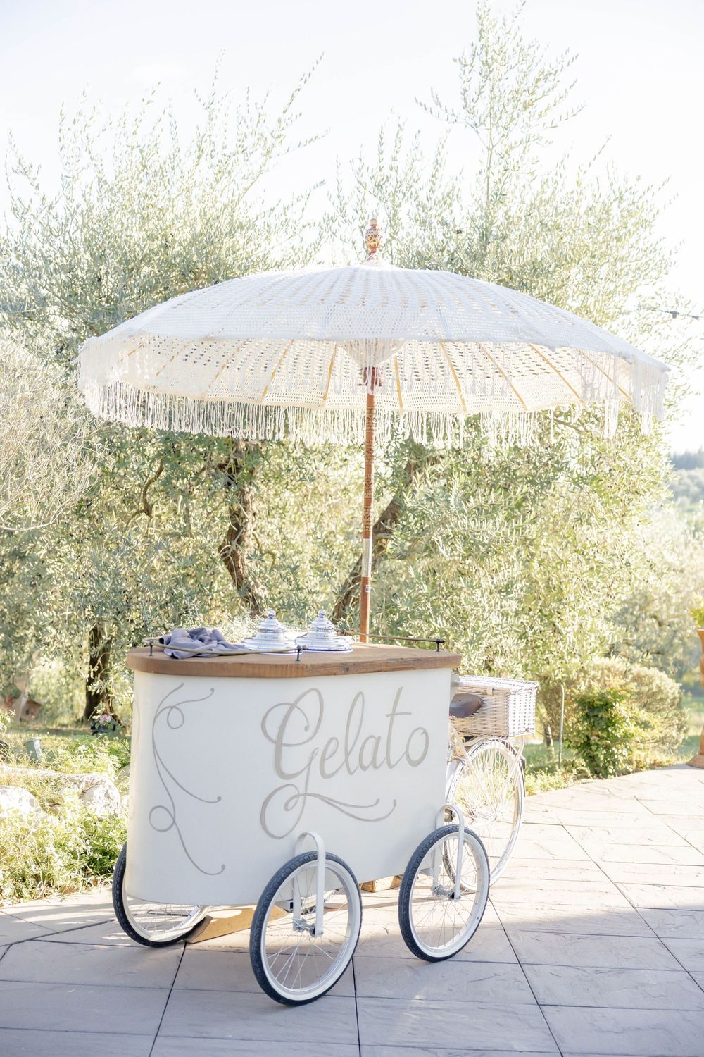 Elegant white gelato cart with fringed umbrella outdoors among olive trees.