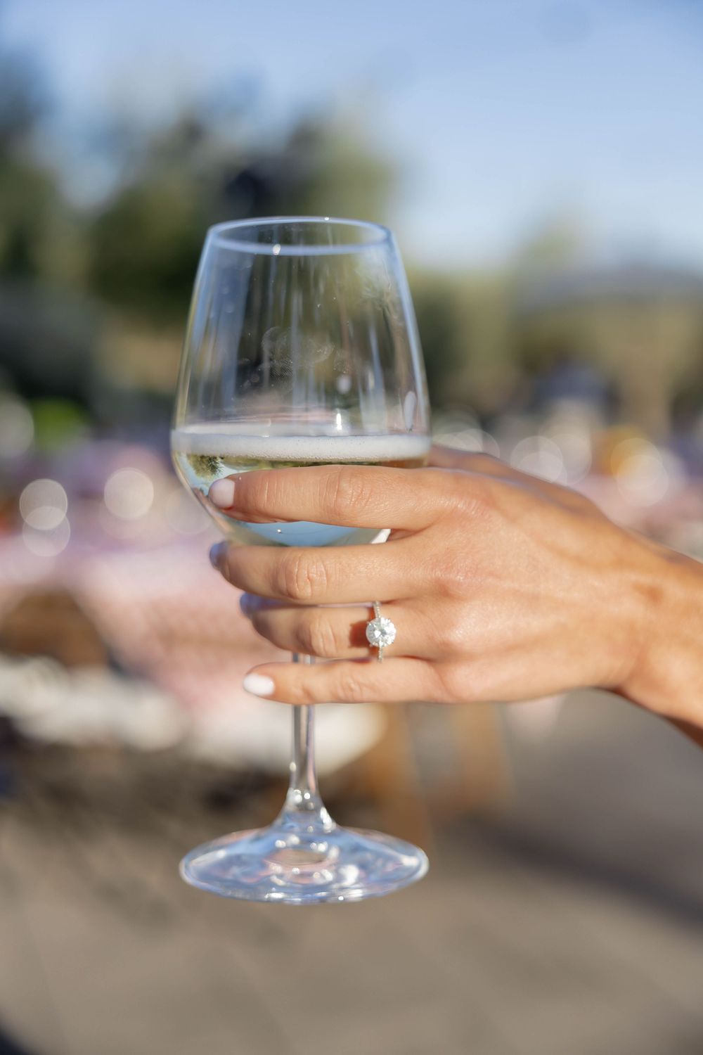 Close-up of a hand wearing a diamond engagement ring while holding a glass of white wine outdoors.