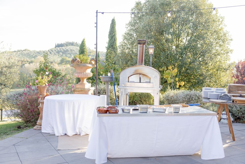 Outdoor pizza oven station with white linen tables set up in a scenic garden venue with cypress trees.