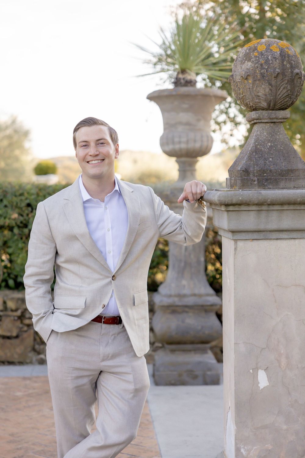 Smiling man in beige suit leaning on stone balustrade in garden.