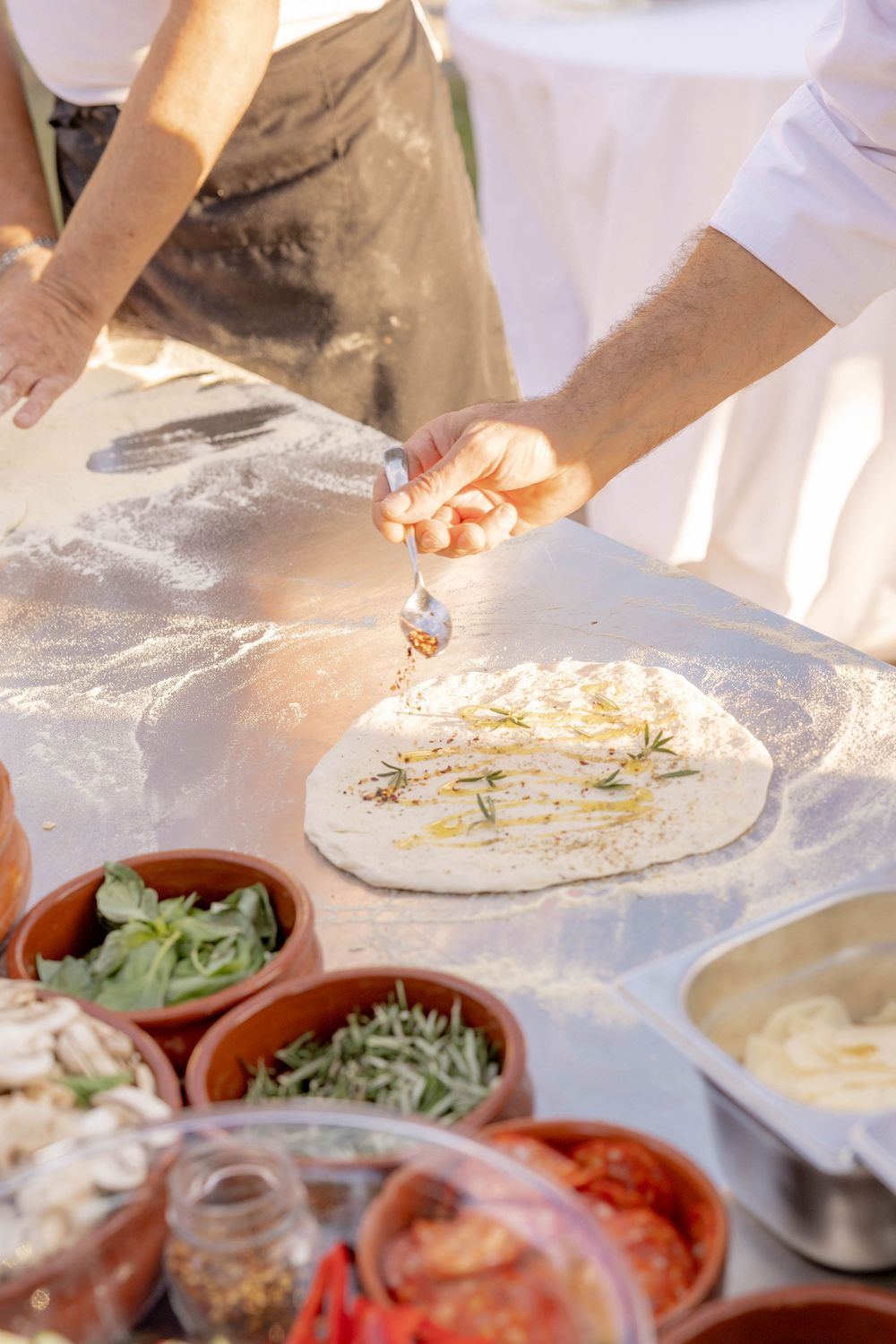 Chef drizzling olive oil on fresh pizza dough with herb toppings and ingredient bowls on a flour-dusted table.
