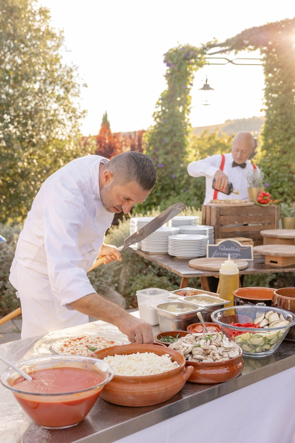 Chef preparing food at an outdoor buffet table with fresh ingredients and a colleague in the background.