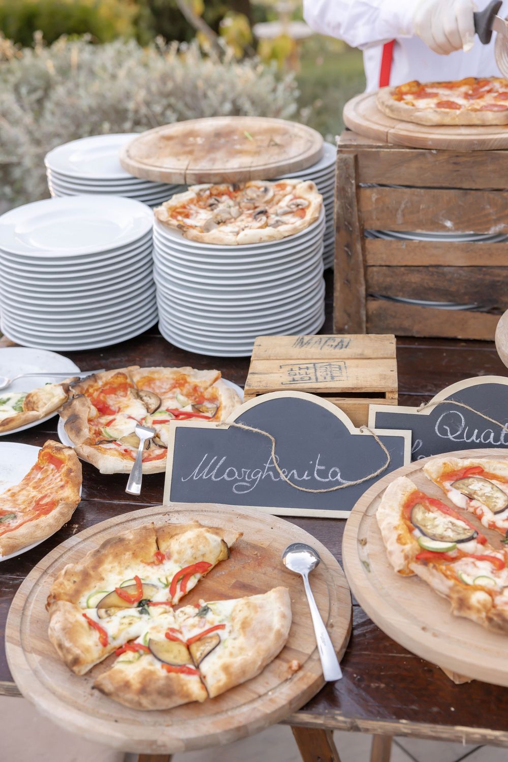Outdoor pizza buffet with Margherita and other varieties on wooden boards, chalkboard signs, and stacked white plates.