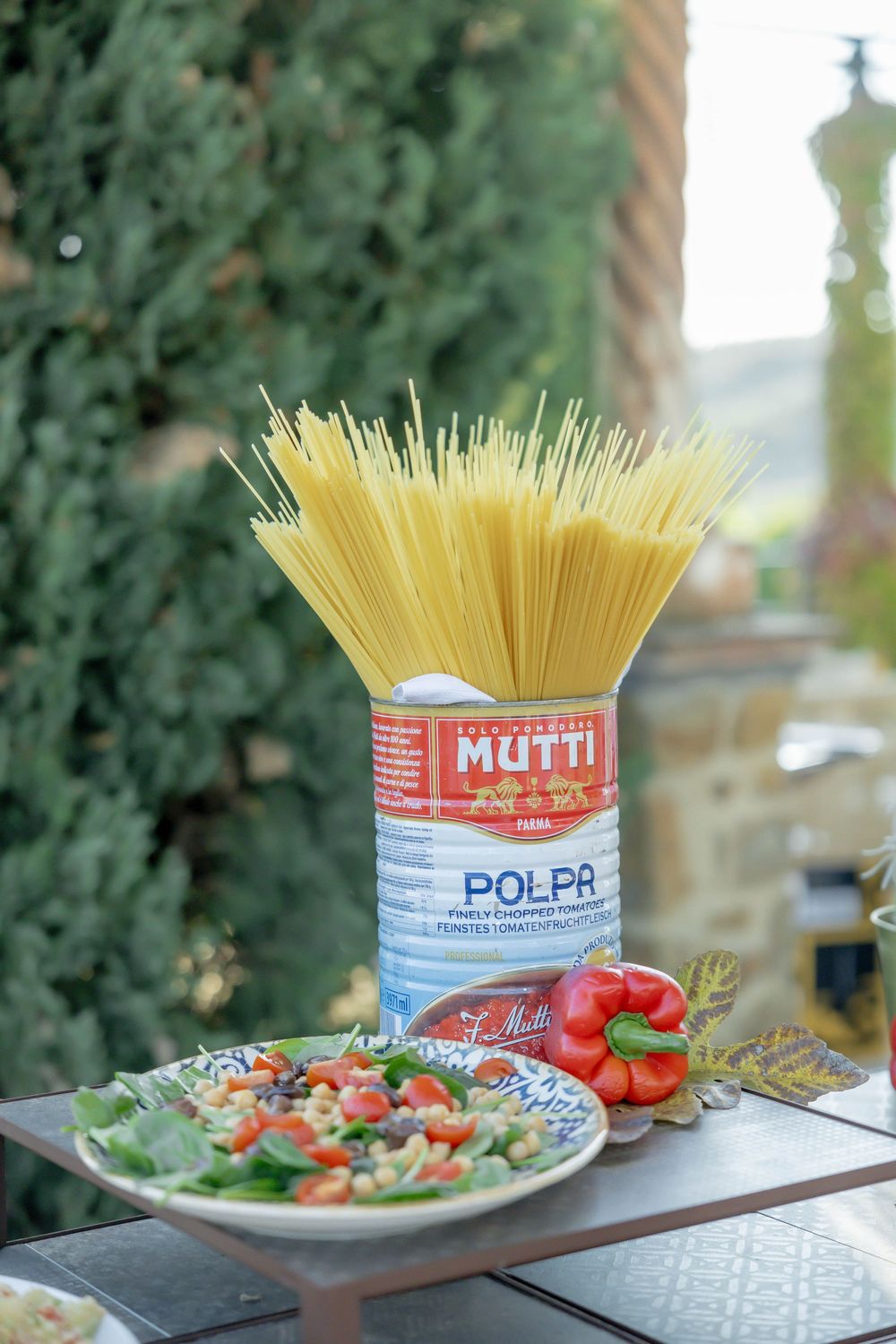 Mutti Polpa can holding dry spaghetti beside a fresh salad with tomatoes and herbs on an outdoor table.