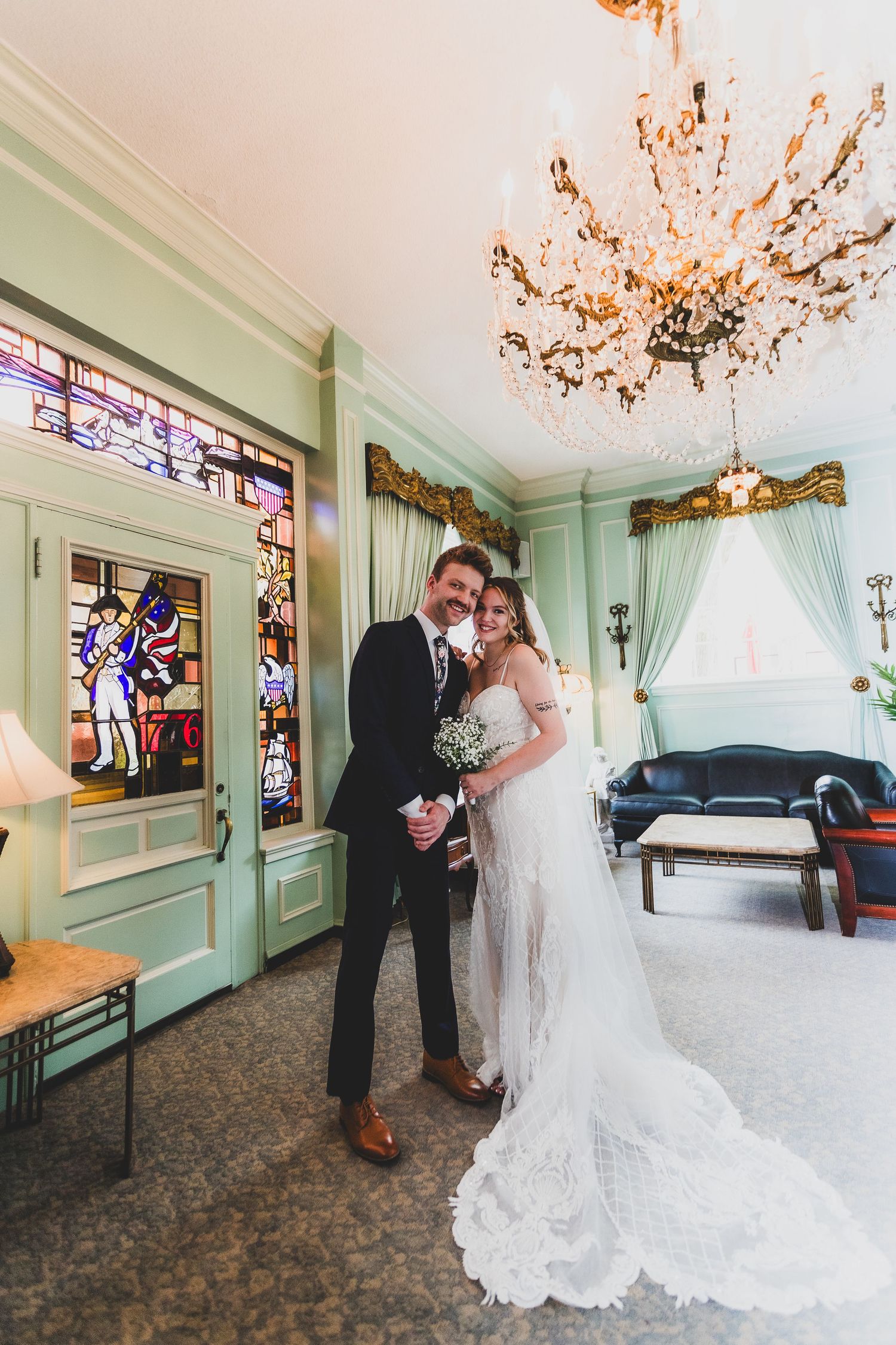 Elegant couple in formal attire posing in vintage room with stained glass windows and crystal chandelier.