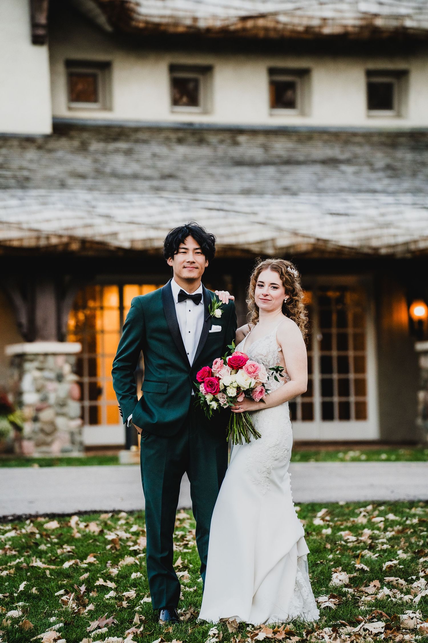 Couple in wedding attire standing in front of rustic cottage with pink bouquet and autumn leaves.