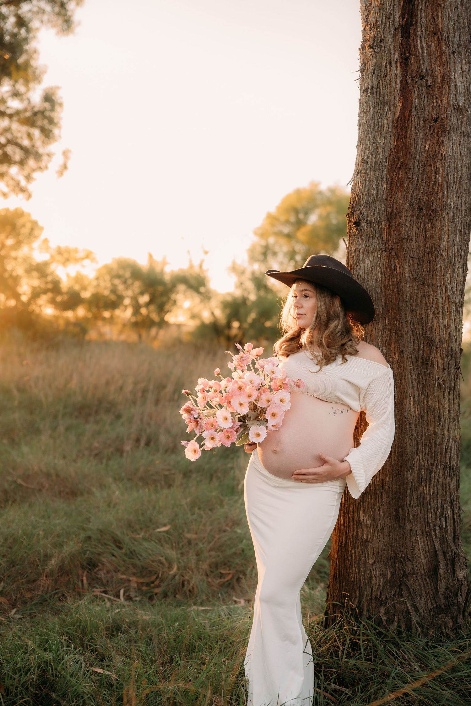 Pregnant woman in white dress and cowboy hat holding pink flowers, leaning against tree at golden hour sunset.