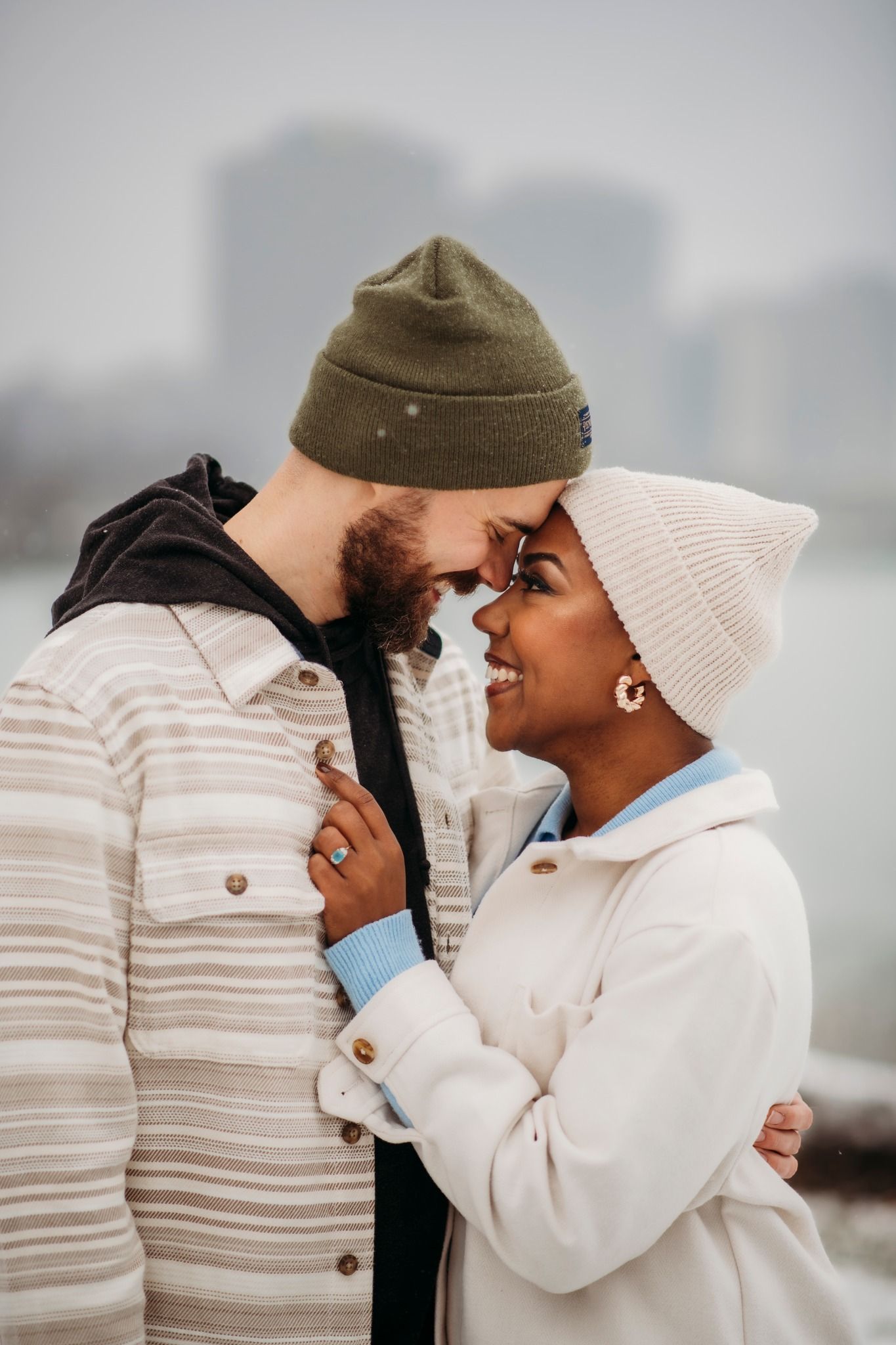 Interracial couple in winter hats sharing a romantic moment outdoors with city skyline in background.