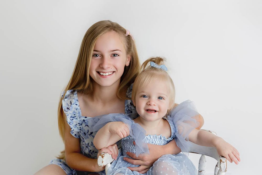 Two sisters in matching blue dresses posing together for a portrait, older girl hugging baby sister.
