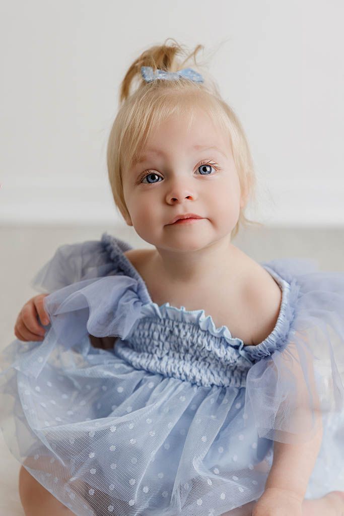 Adorable blonde toddler girl in a blue tulle dress with a bow hair clip poses for a portrait photo.