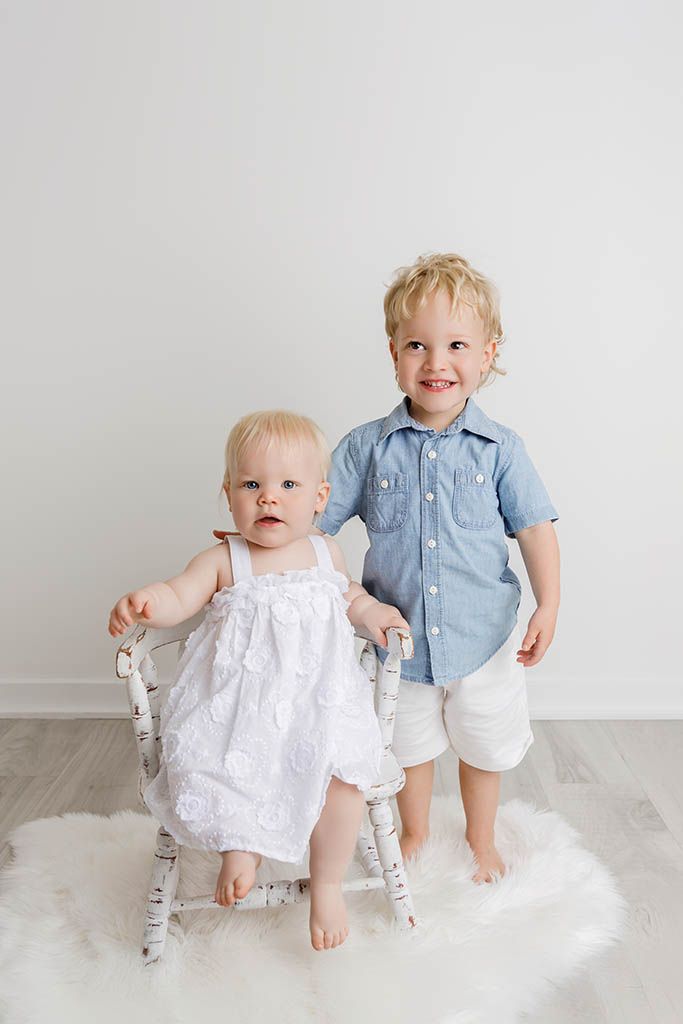 Two young siblings, a baby girl in white dress and toddler boy in blue shirt, pose together for a portrait.