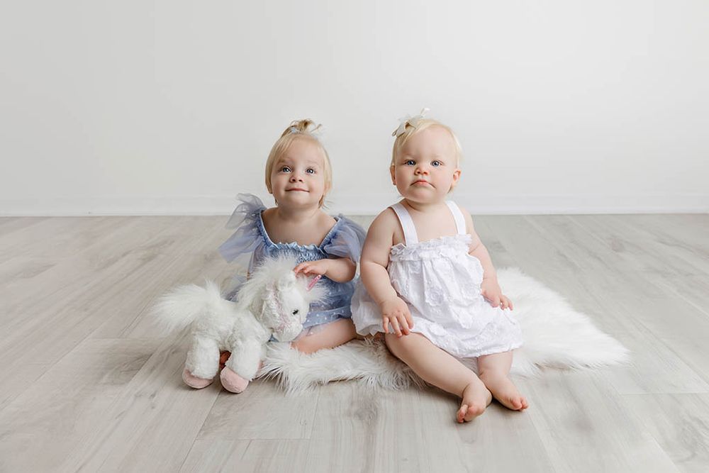 Two baby girls in pastel dresses sit on a fur rug by Canyon Lake infant photographer.