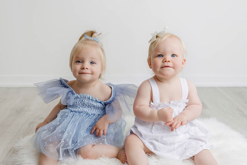 Two adorable baby girls in blue and white dresses sitting together on a white fluffy rug by Canyon Lake Photographer.