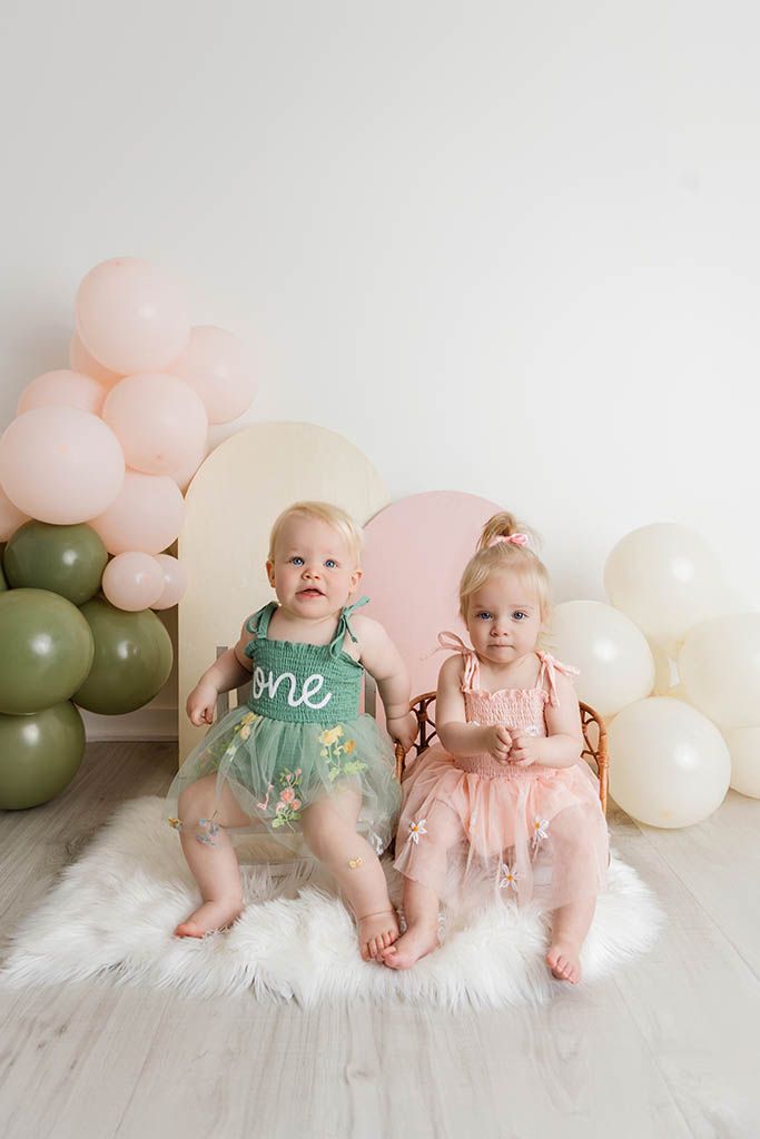 Two baby girls in green and pink tutu dresses sit on fur rug surrounded by pastel balloons.