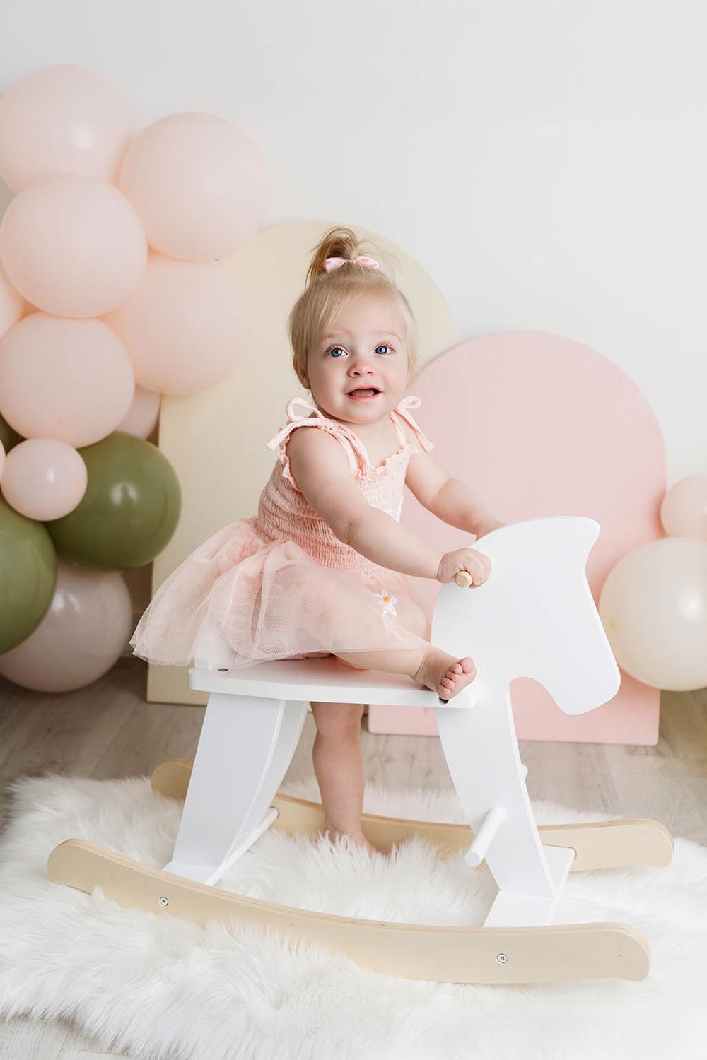 Smiling baby girl in pink tutu dress stands on white wooden rocking horse with balloons by Canyon Lake baby's first birthday photographer.