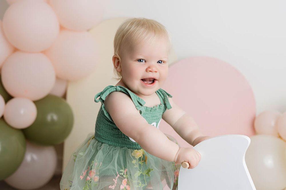 Happy baby girl in green floral dress standing by white chair with pink balloon backdrop by professional Canyon Lake baby photographer.