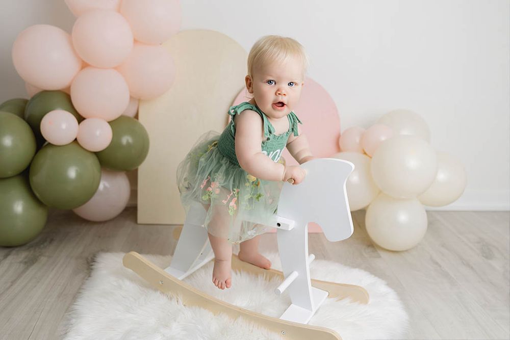 Canyon Lake Photography - Baby girl in green floral tutu dress stands on white wooden rocking horse amid pastel balloons.