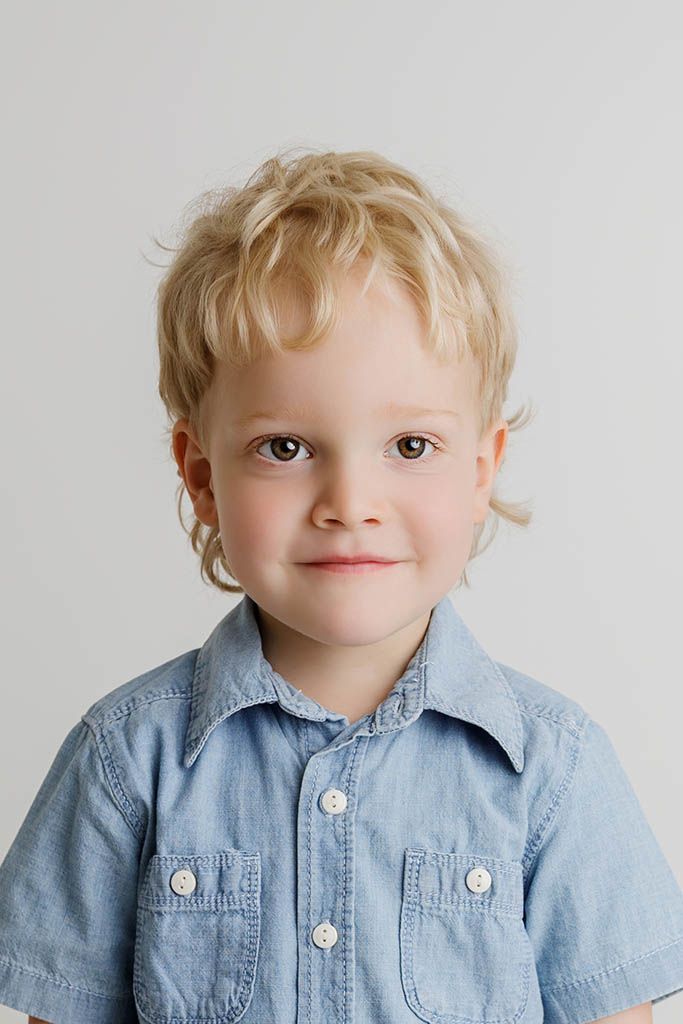 Cute blonde toddler boy with curly hair smiling in a light blue denim button-up shirt.