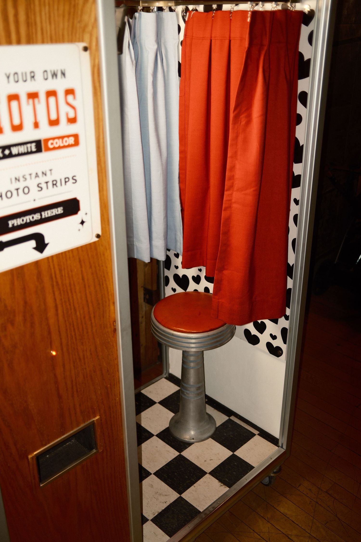 Vintage photo booth with red curtain, cow print walls, checkered floor, and chrome stool offering black & white or color photo strips.