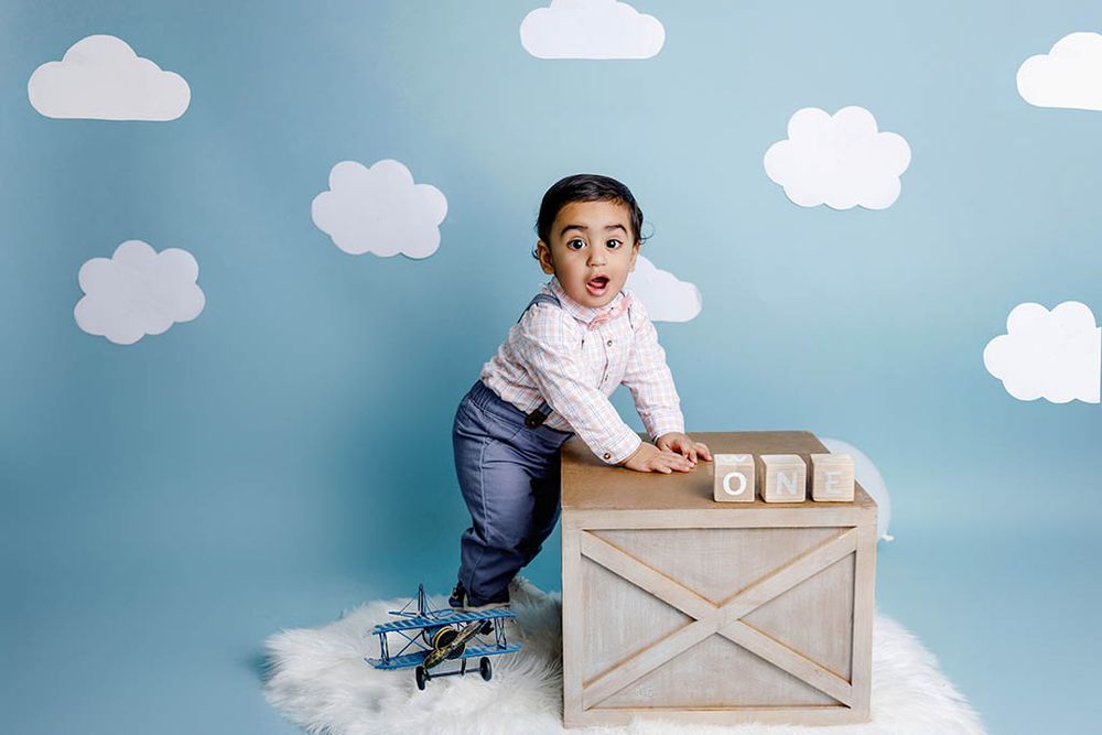 Adorable toddler boy posing with wooden blocks and toy airplane on cloud-themed blue backdrop.