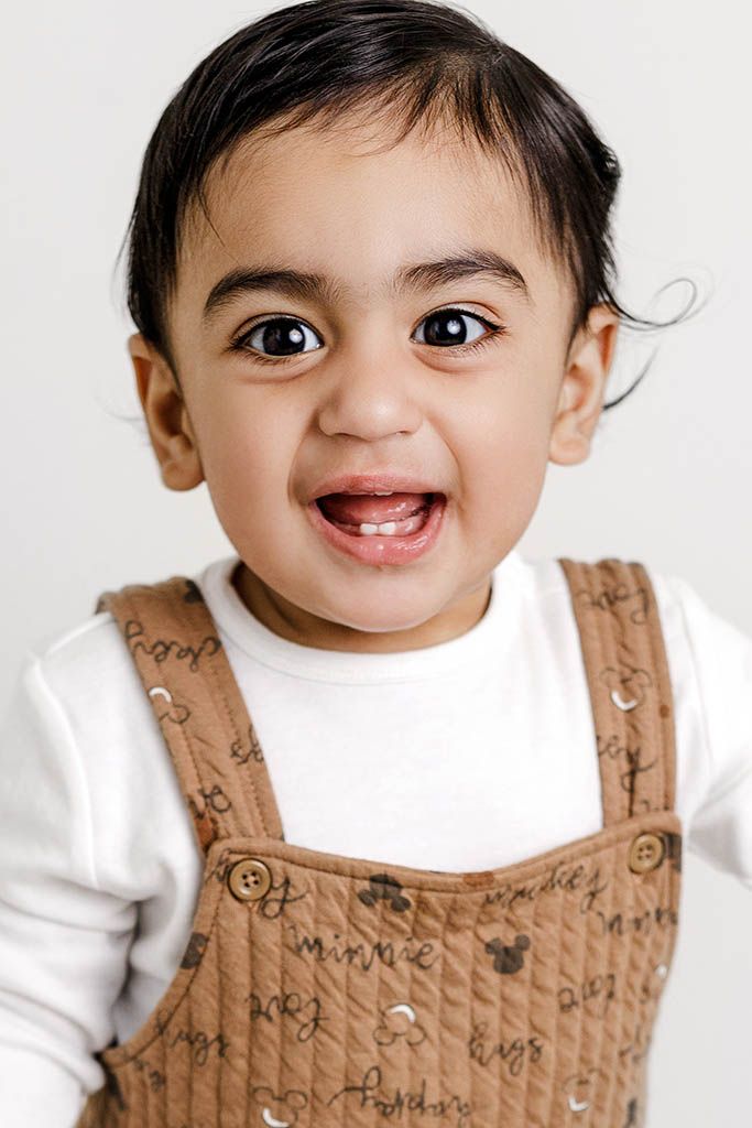 Portrait by Moreno Valley baby photographer: Smiling baby boy wearing a brown Mickey Mouse overalls and white long-sleeve shirt.