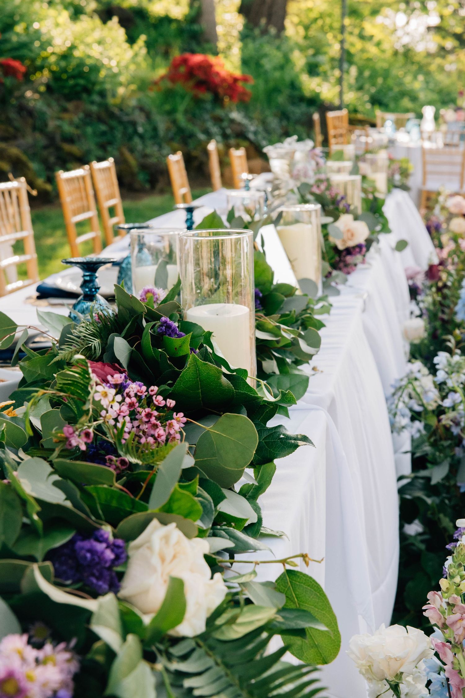 Elegant outdoor wedding reception table adorned with lush greenery garland, white candles, and purple and pink flowers.