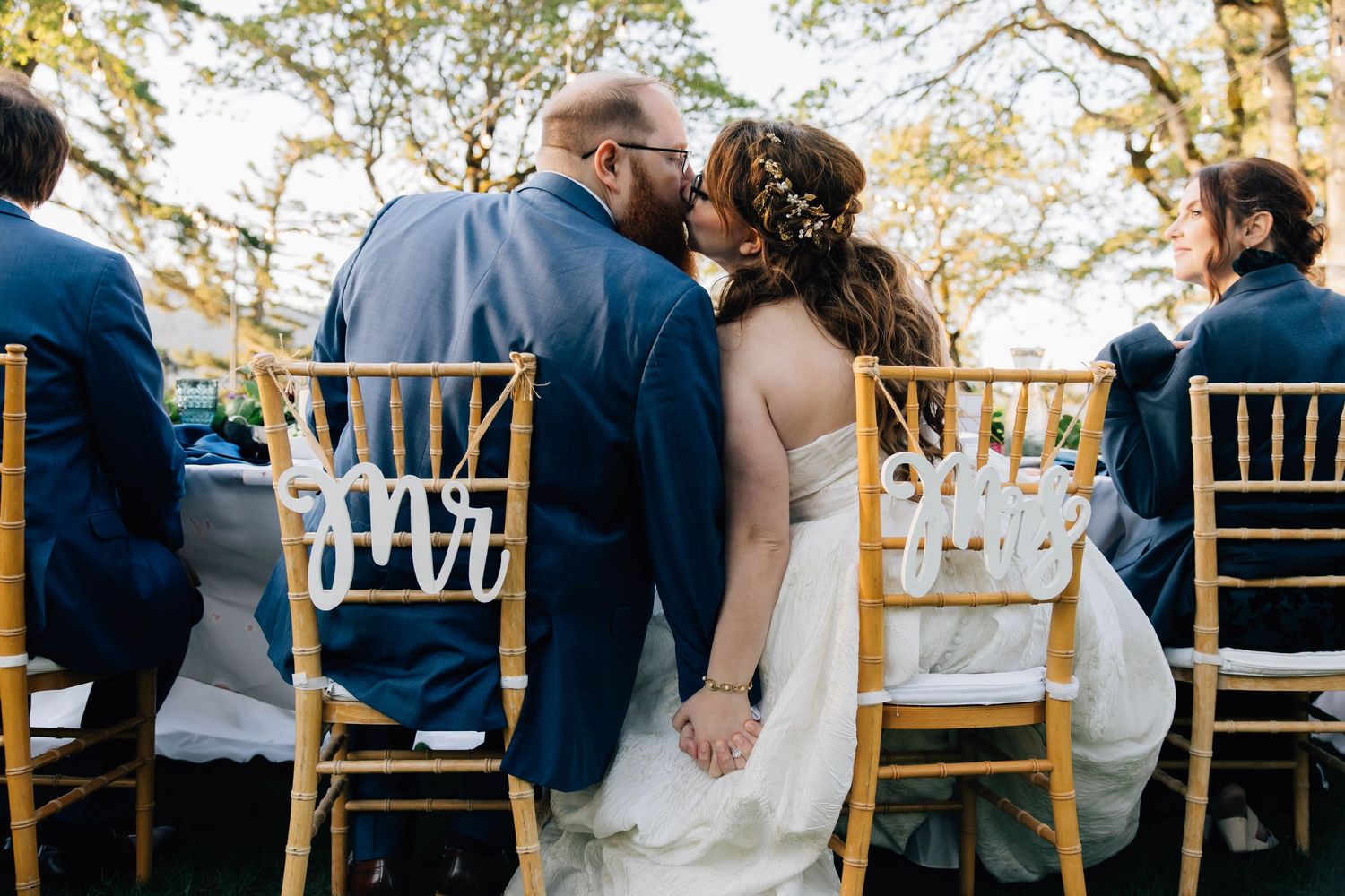 Bride and groom kissing at outdoor wedding ceremony, seated in gold chiavari chairs with Mr and Mrs signs.